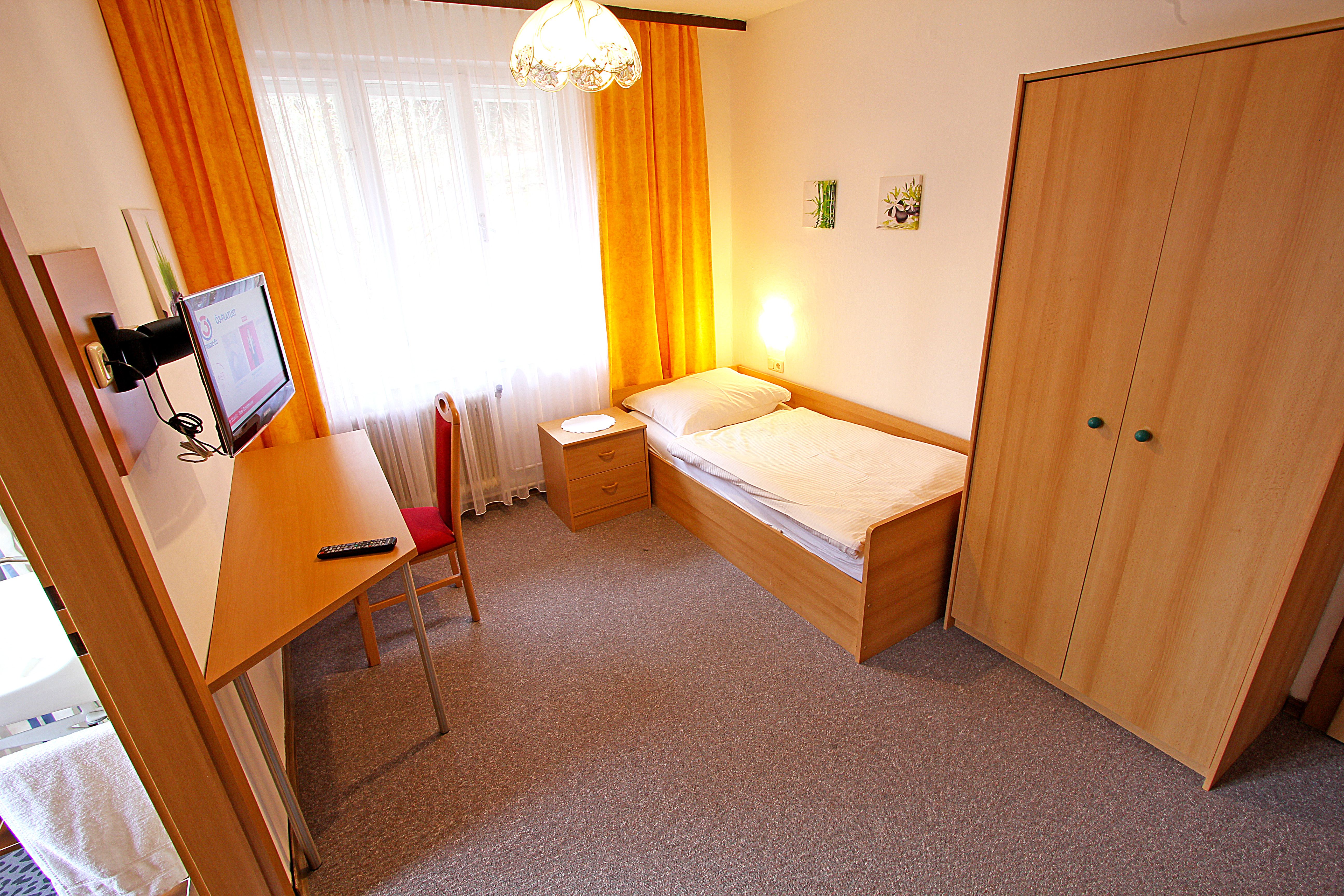 Ein einfach eingerichtetes Einzelzimmer mit Bett, Schreibtisch, Stuhl, Kleiderschrank und Fernseher. Gelbe Vorhänge und Teppichboden.
