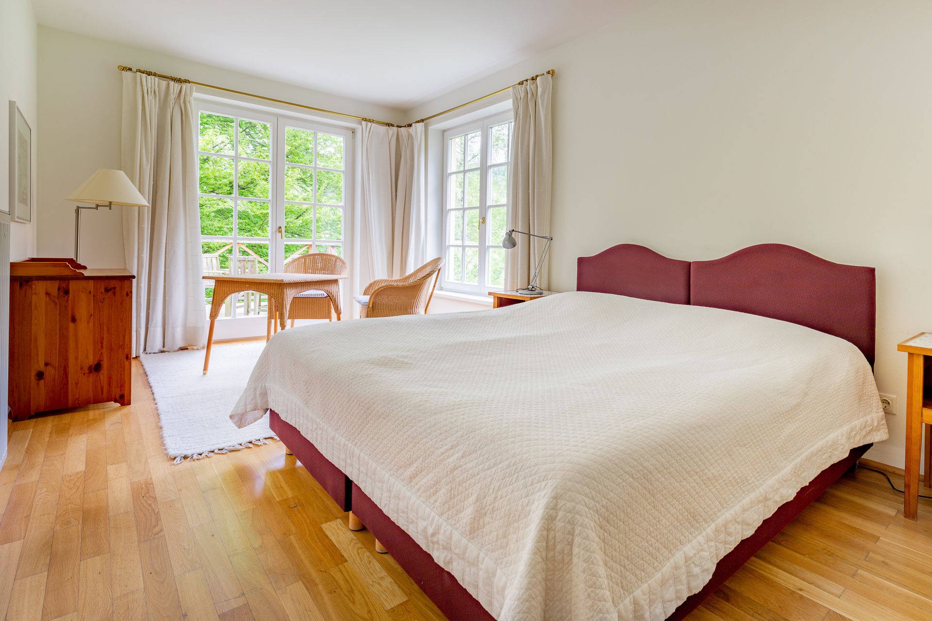 Helles Schlafzimmer mit großem Bett, Holzboden, zwei Korbstühlen und Tisch vor großen Fenstern mit Blick ins Grüne.