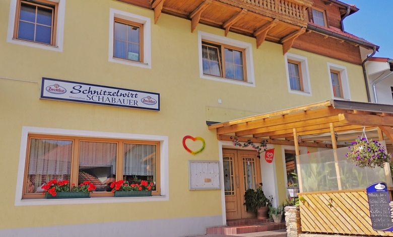 Gelbes Gebäude mit Schild 'Schnitzelwirt Schabauer', Blumenkästen und überdachtem Eingang.