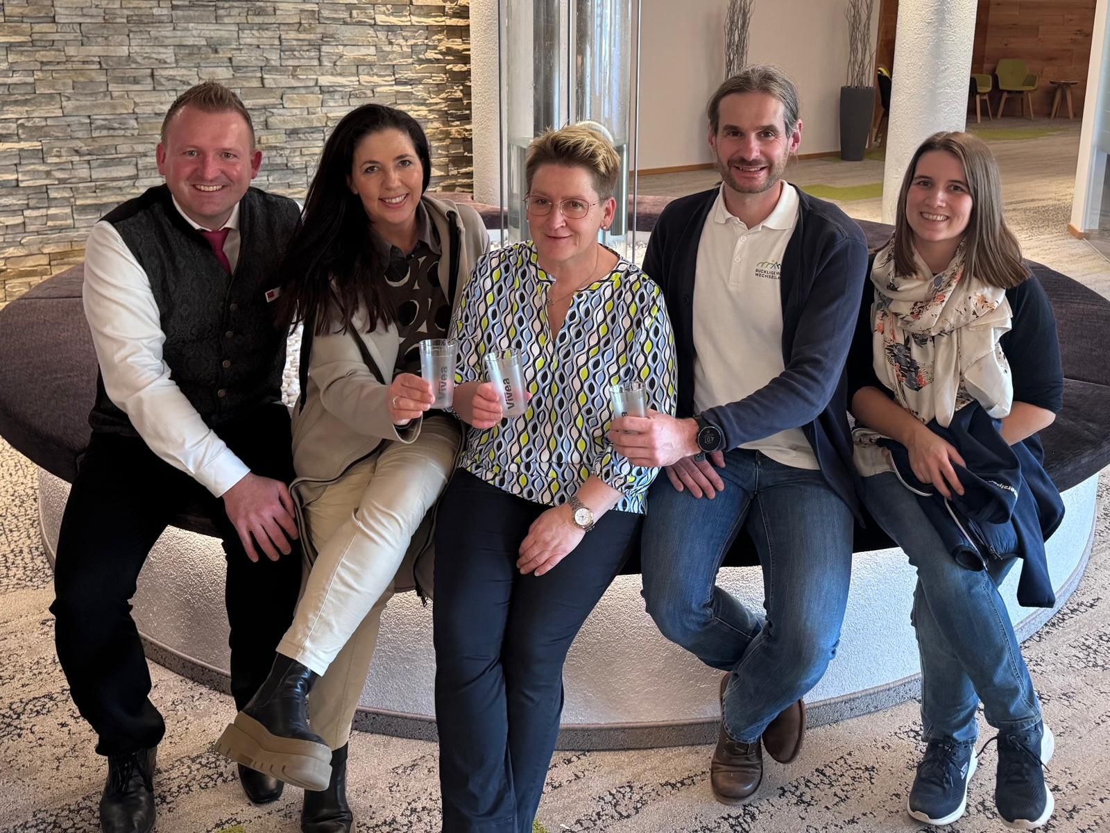 Fünf Personen sitzen auf einer Bank, drei Personen davon halten einen Becher in den Händen.