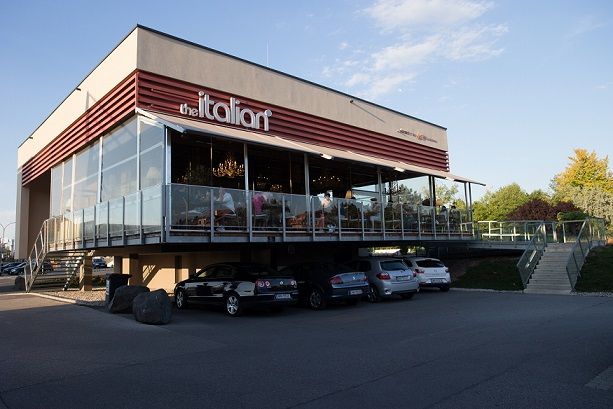 Modernes Restaurant mit Glasfassade und Terrasse, Autos parken darunter.
