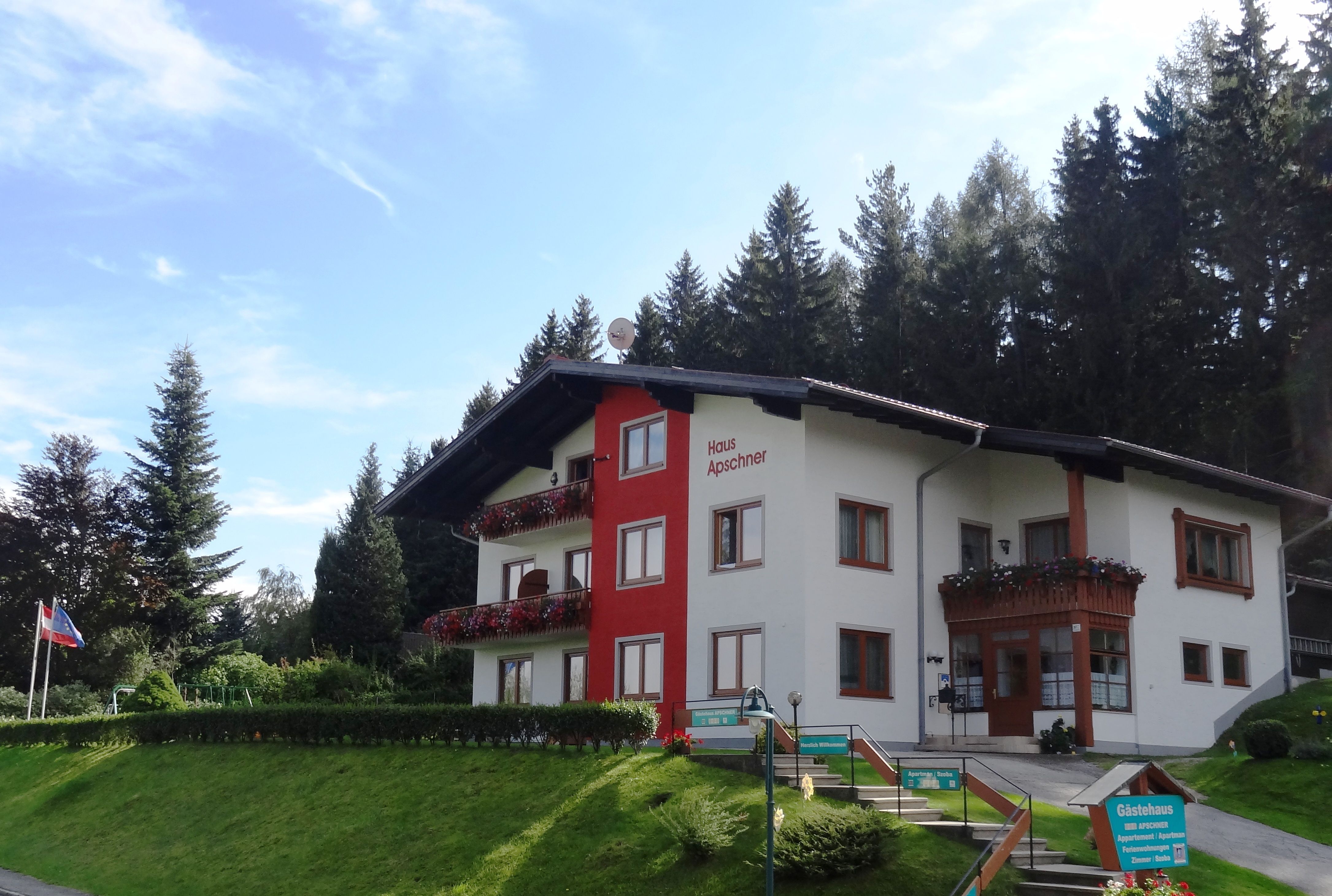 Ein zweistöckiges Gästehaus mit roten Balkonen und Blumen, umgeben von Bäumen und einem blauen Himmel.