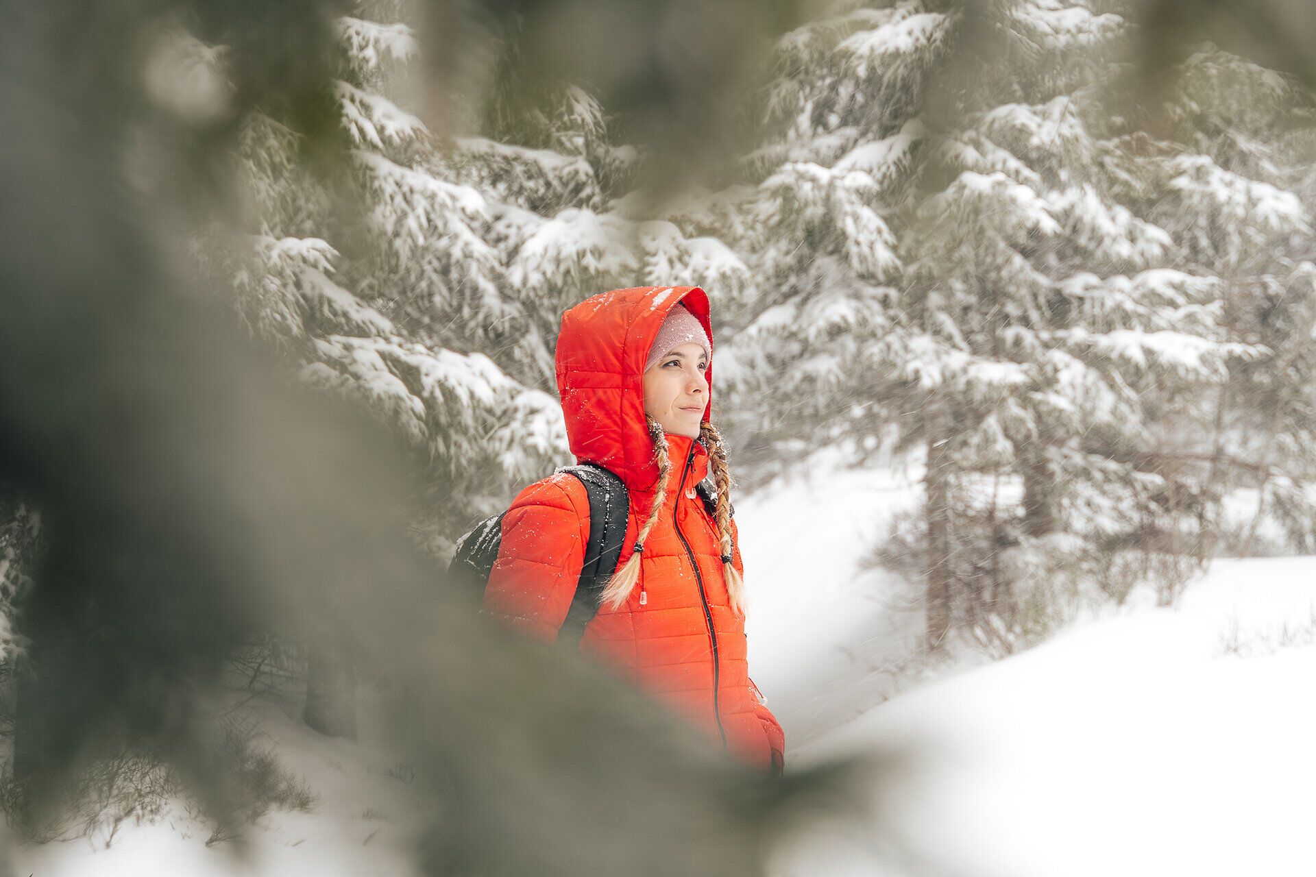Inmitten einer winterlichen Märchenlandschaft strahlt die leuchtend rote Jacke der Wanderin gegen den schneebedeckten Wald. Die sanften Flocken fallen leise und verleihen der Umgebung eine friedliche Stille, während die frische Bergluft ein Gefühl von Freiheit und Abenteuer vermittelt.