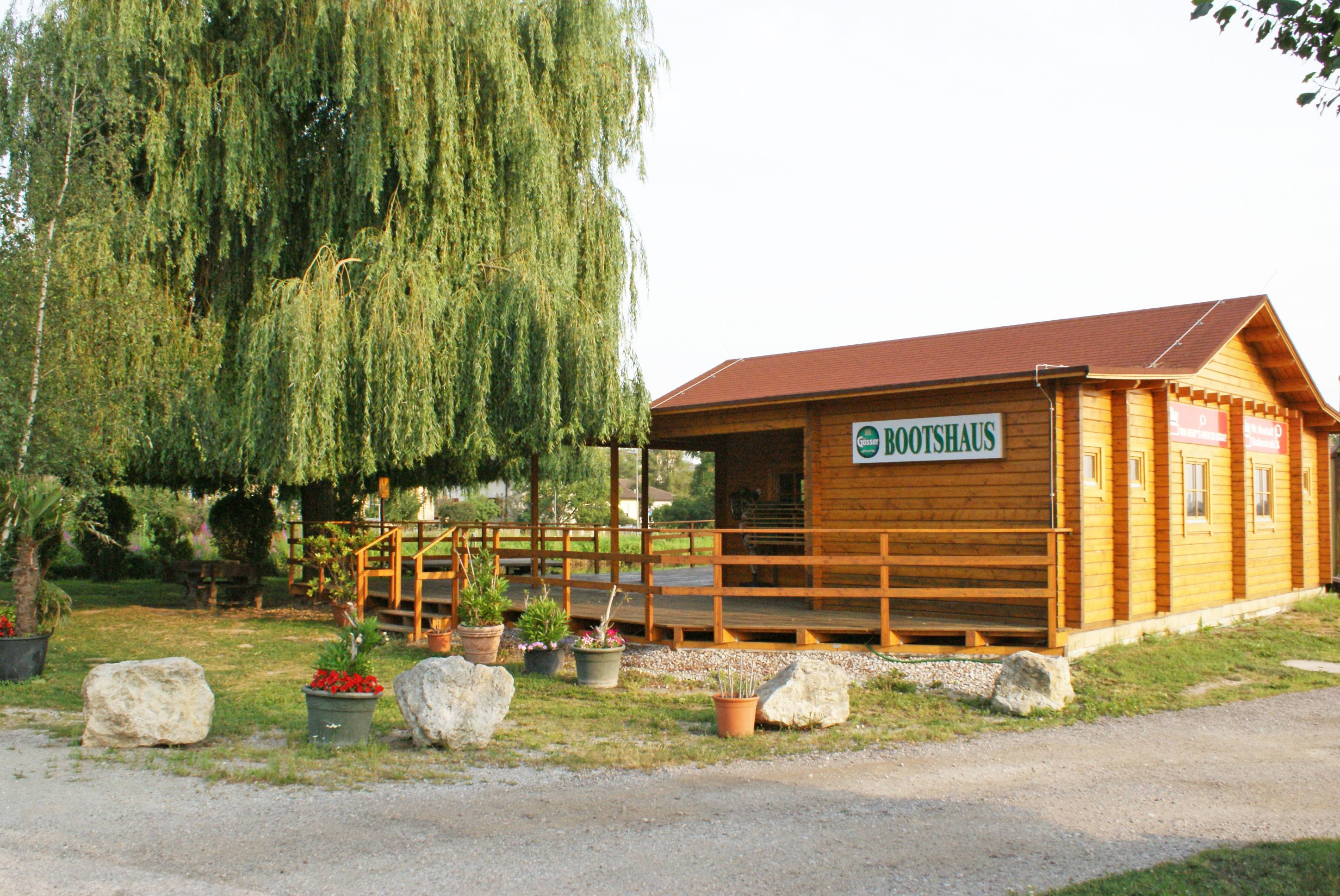 Holzgebäude mit Schild 'Bootshaus' neben einem großen Baum und Pflanzen.