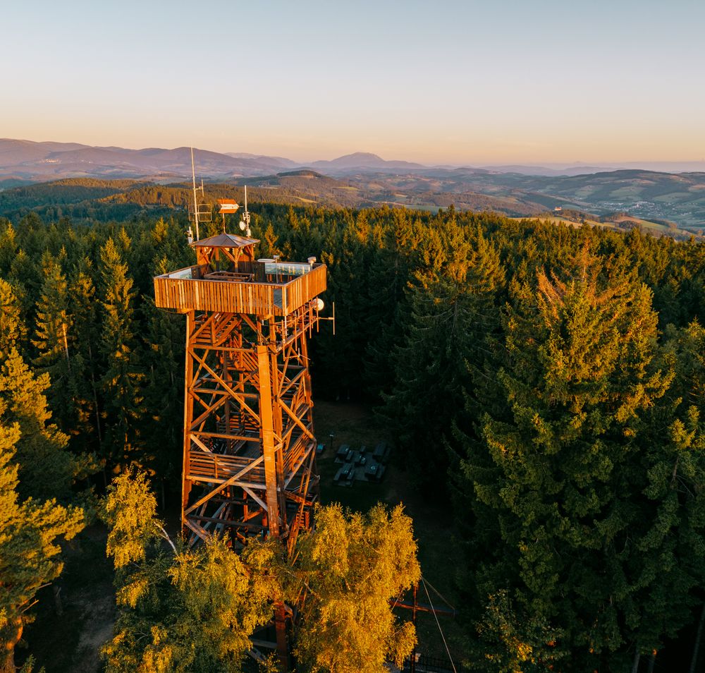 Aussichtsturm aus Holz im Wald mit zwei Personen, die den Sonnenuntergang betrachten, im Hintergrund hügeliges Panorama,