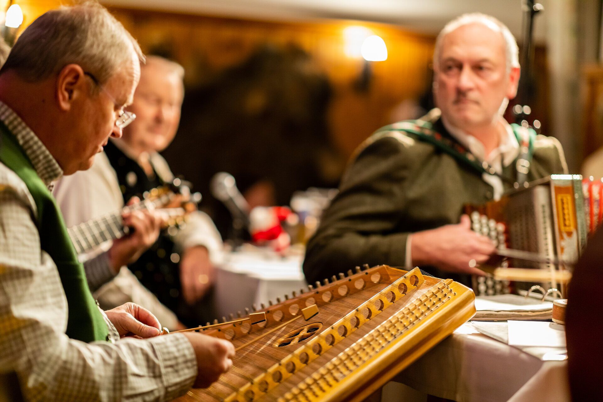 Männer musizieren gemeinsam mit Zither, Akkordeon und Gitarre in gemütlicher Stubenatmosphäre.