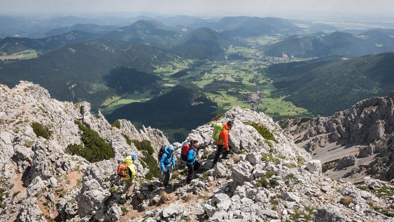 Group of hikers on a rocky mountain path with a view of a green valley plain and surrounding mountains.