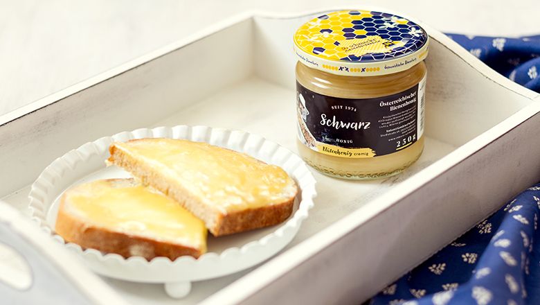 Ein weißes Holztablett mit einem Glas Honig und einem weißen Teller mit zwei Scheiben Honigbrot.