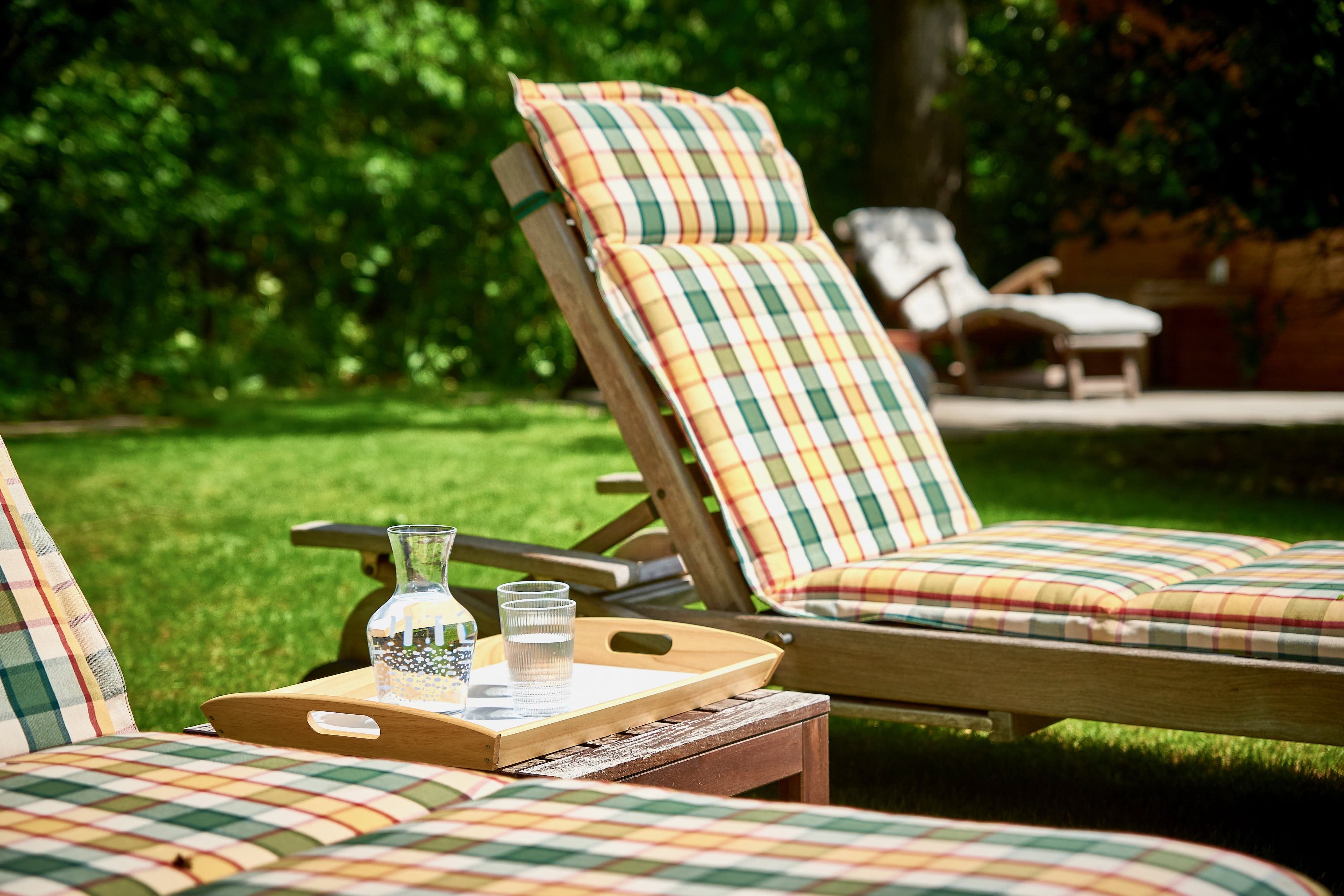 Sonnenliegen mit karierten Auflagen und einem Tablett mit Wasserkrug und Glas im Garten.