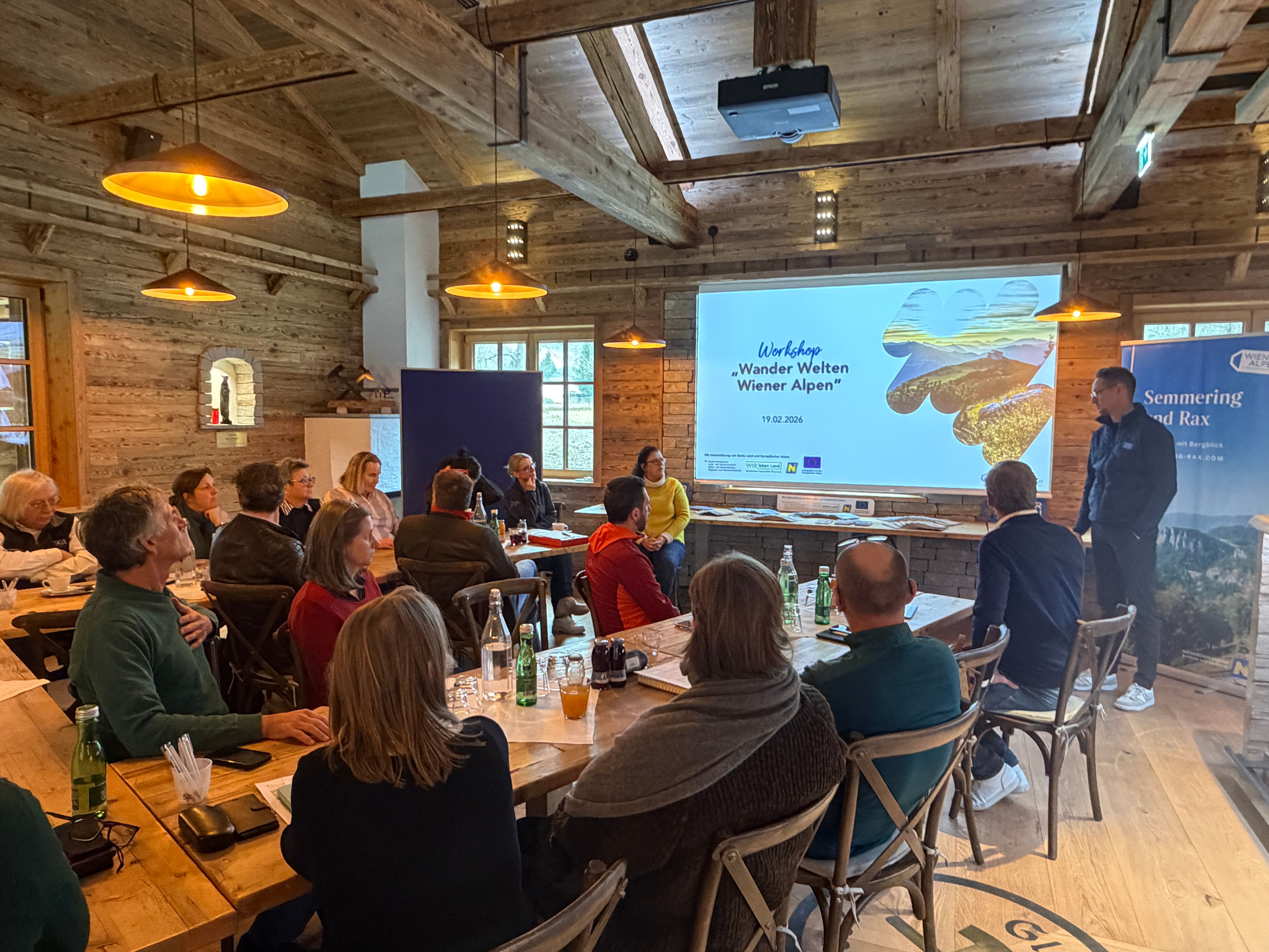 Workshop Teilnehmer sitzen in einem rustikalen Raum an Tischen, ein Mann moderiert den Workshop vor den Teilnehmern vor einer Leinwand. 