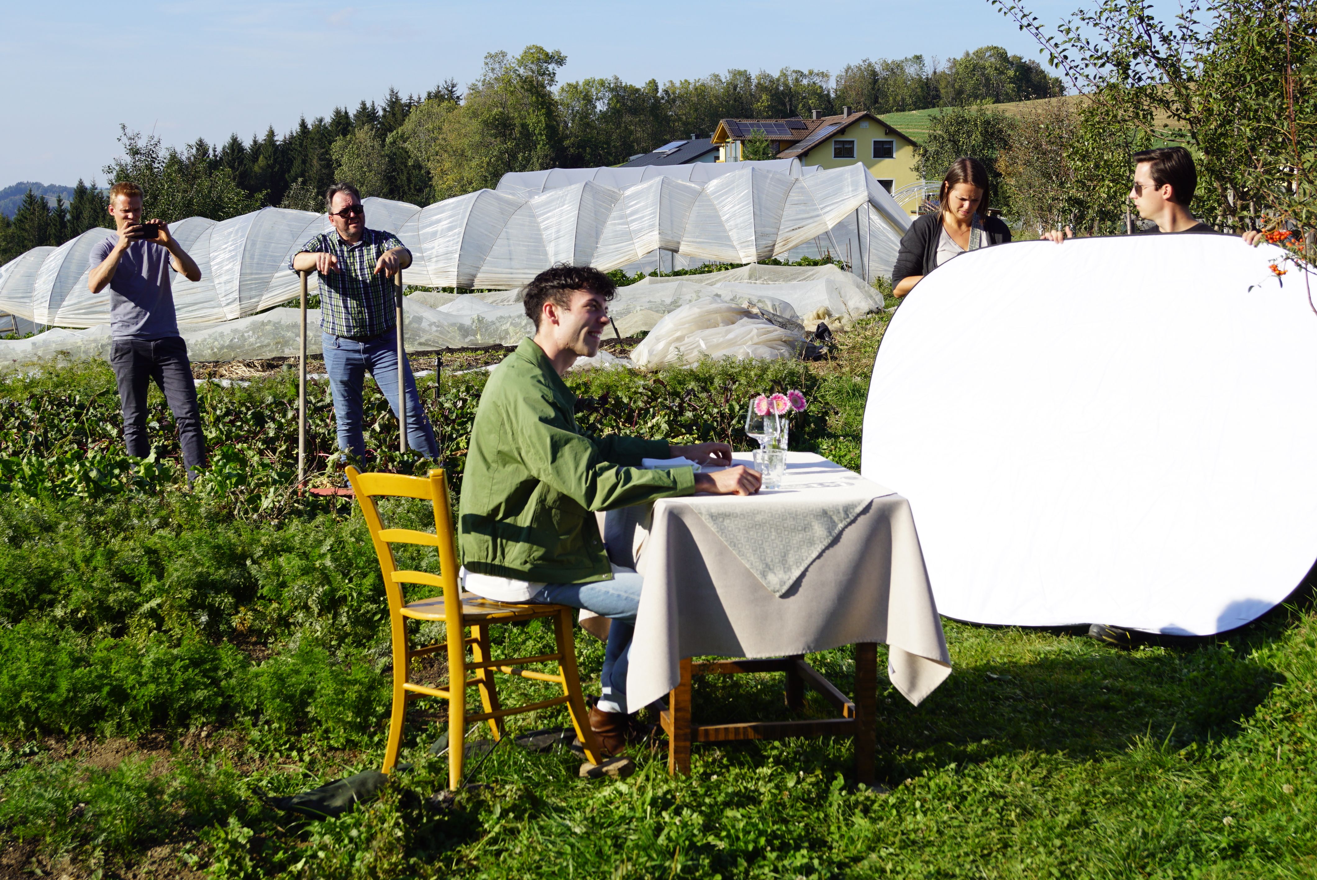 Ein junger Mann sitzt auf einem Feld bei einem gedeckten Tisch, rundherum stehen Foto- und Videoproduzenten mit Equipment und weitere Darsteller.