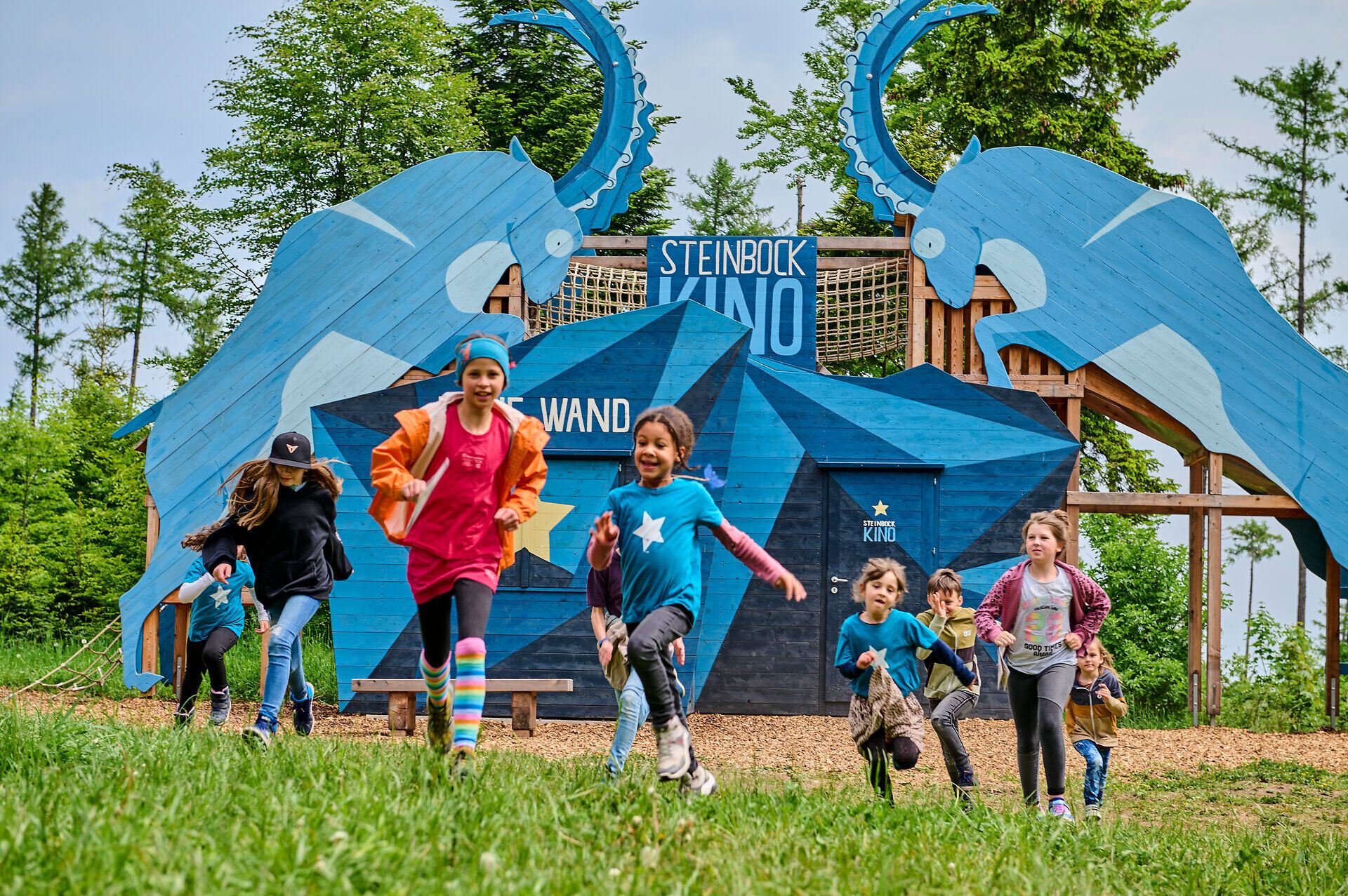 Eine Gruppe fröhlicher Kinder läuft voller Begeisterung über die Wiese, umgeben von der beeindruckenden Kulisse der Natur. Die bunten Farben ihrer Kleidung spiegeln die Lebhaftigkeit des Sommers wider, während die majestätischen Figuren im Hintergrund die Abenteuerlust anregen.