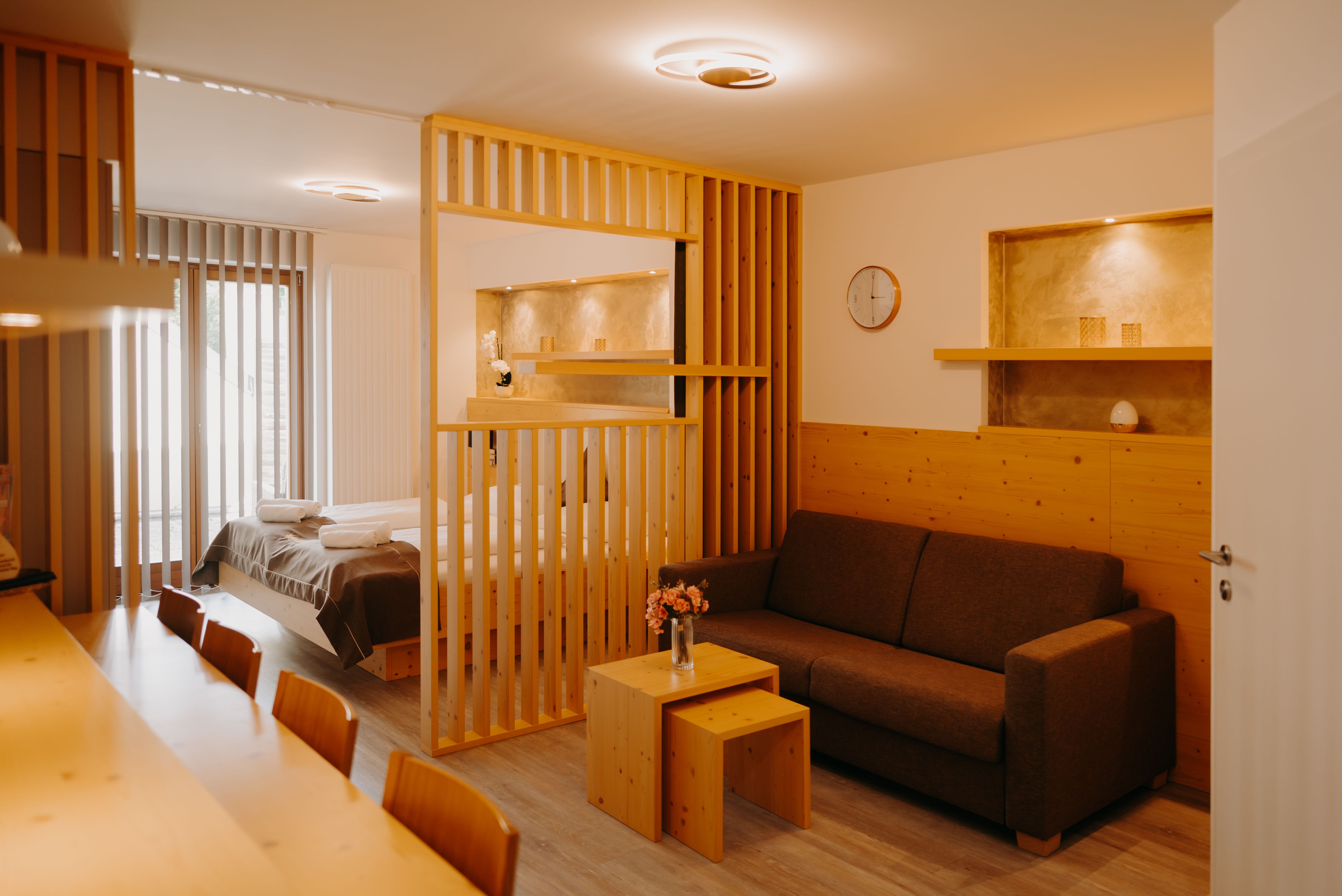 Modernes Hotelappartement mit Doppelbett, hellen Holzmöbeln, ausziehbarer Couch und Raumtrenner aus Holz. Die Wand ist zum Teil mit Holz verkleidet und in der Wand sind Nischen, die in einem erdigen Gold gestrichen sind.