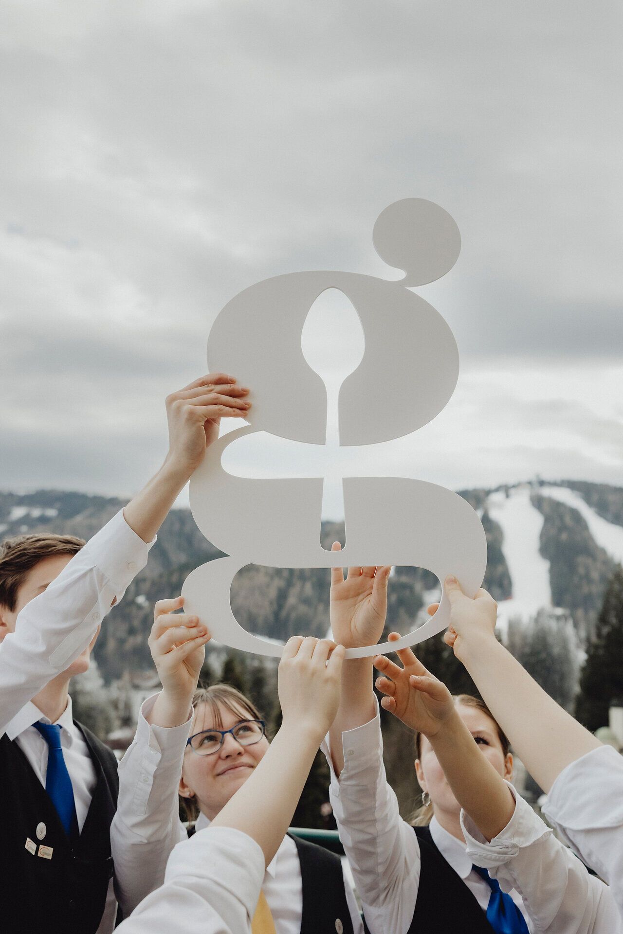 Gruppe junger Menschen hebt gemeinsam ein ausgeschnittenes Symbol vor einer Berglandschaft in die Höhe.
