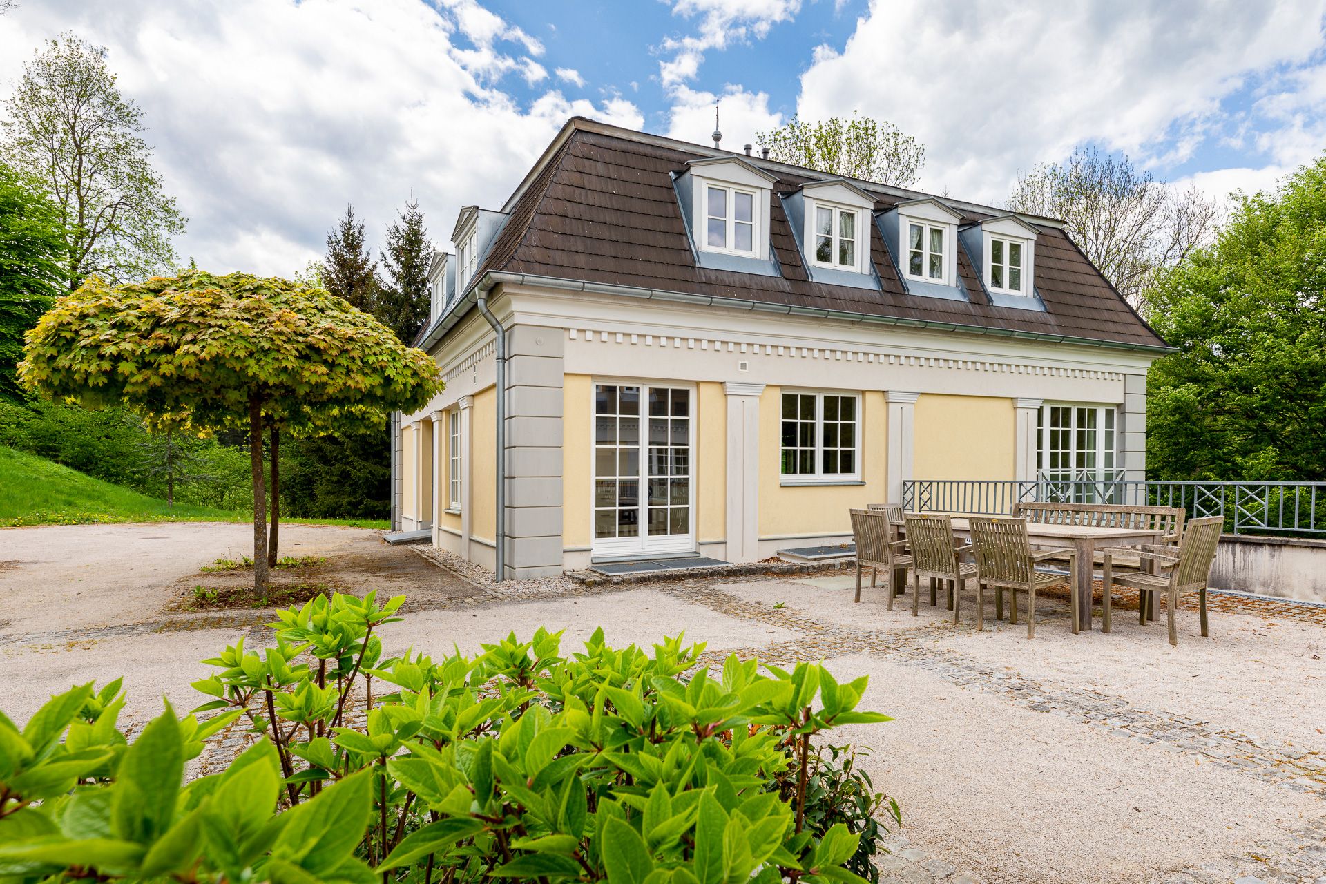 Gelbes Kavaliershaus mit Mansardendach, umgeben von Bäumen und Gartenmöbeln im Vordergrund.
