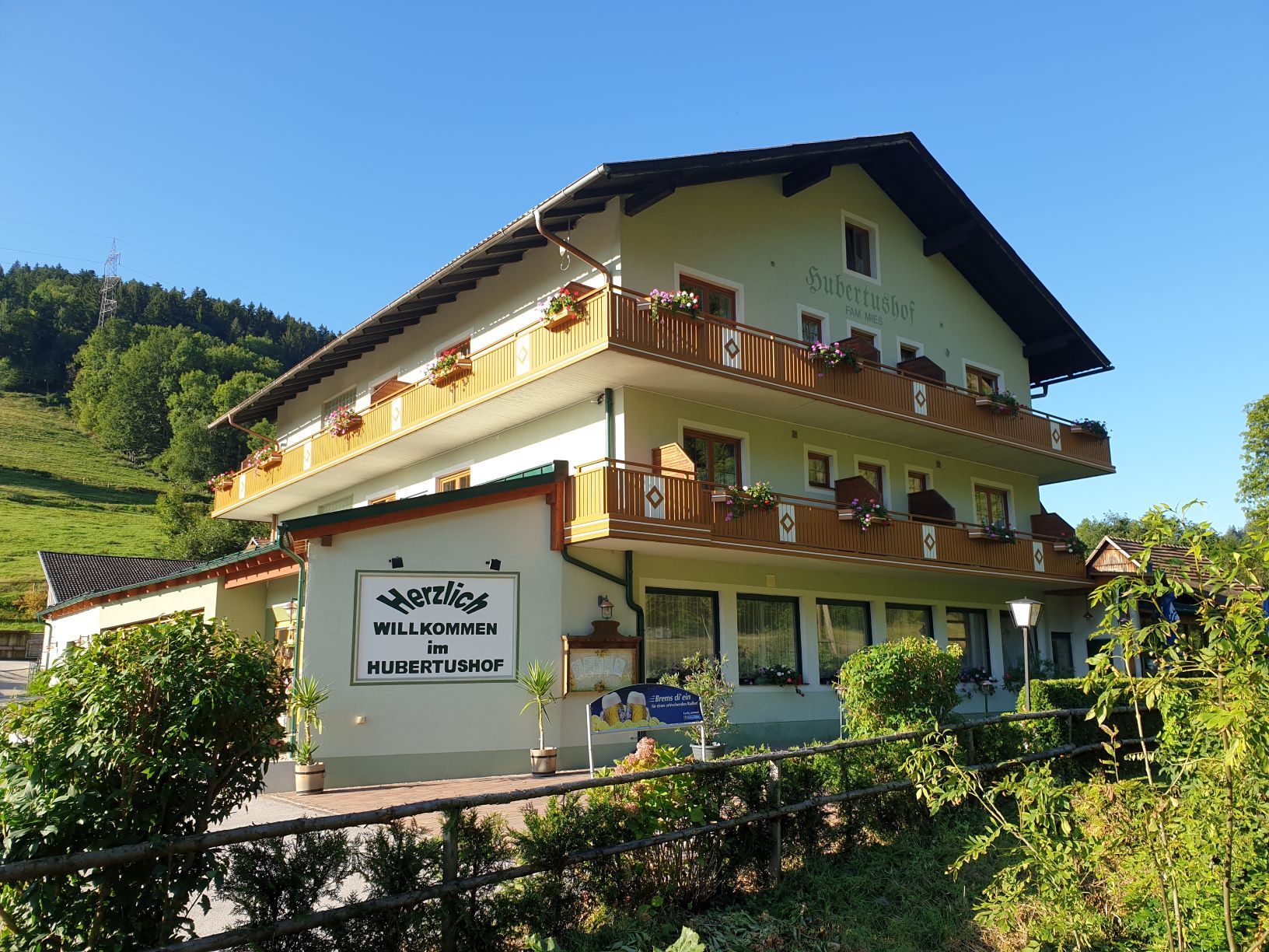 Ein traditionelles Gasthaus namens Hubertushof mit Blumen auf den Balkonen, umgeben von grüner Landschaft und blauem Himmel.