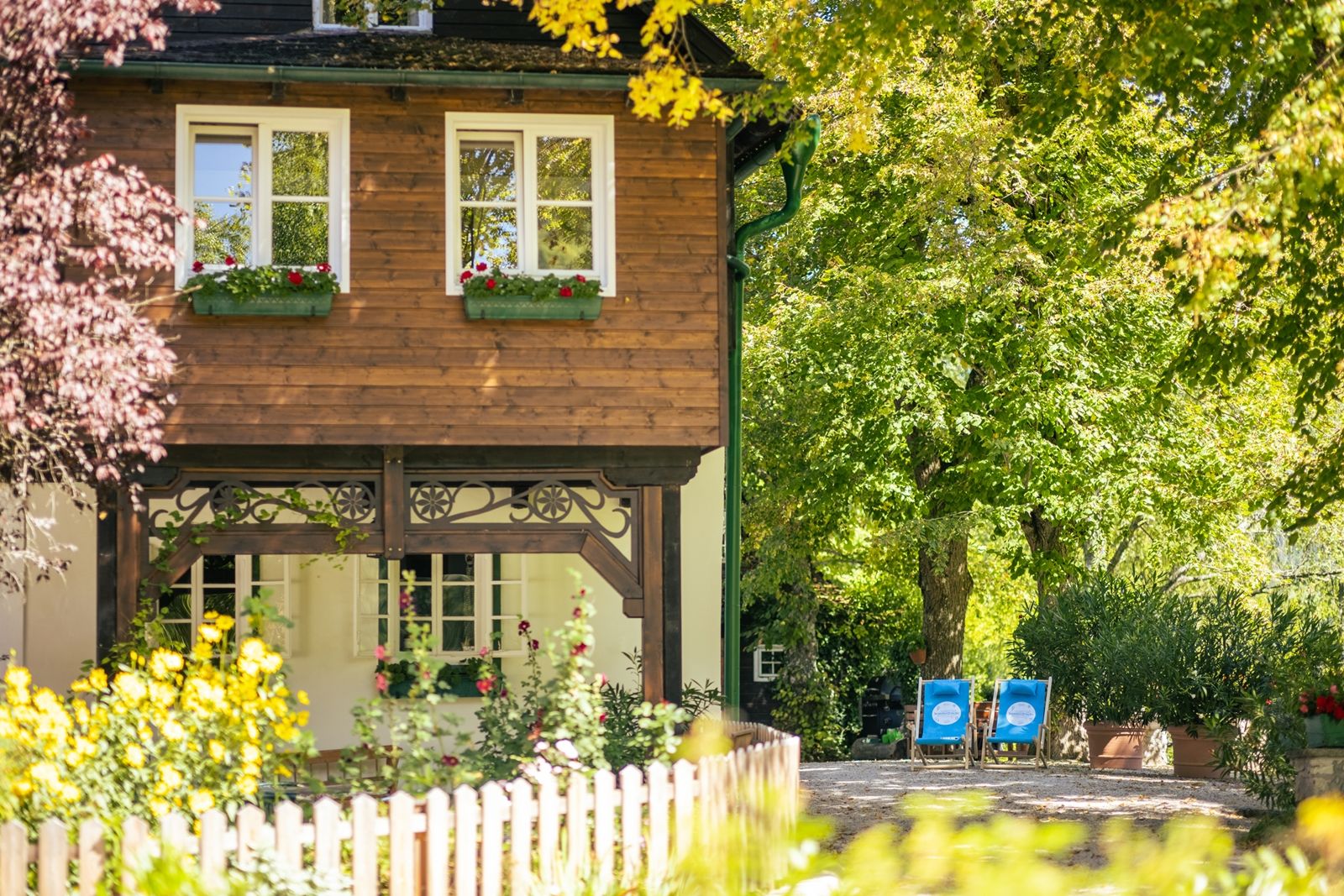 Ein idyllisches Landhaus mit Holzelementen, umgeben von blühenden Gärten und Bäumen. Zwei blaue Liegestühle stehen im Garten.
