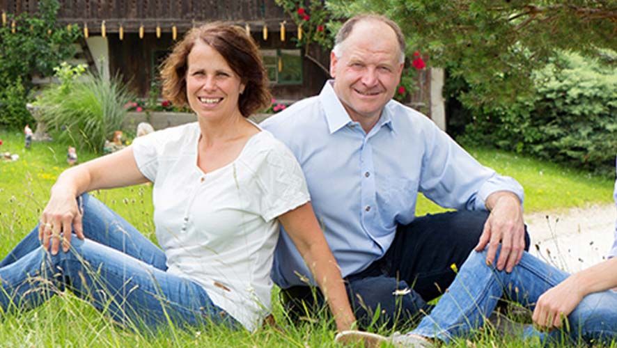 Ein Mann und eine Frau sitzen lächelnd auf einer Wiese vor einem Haus.
