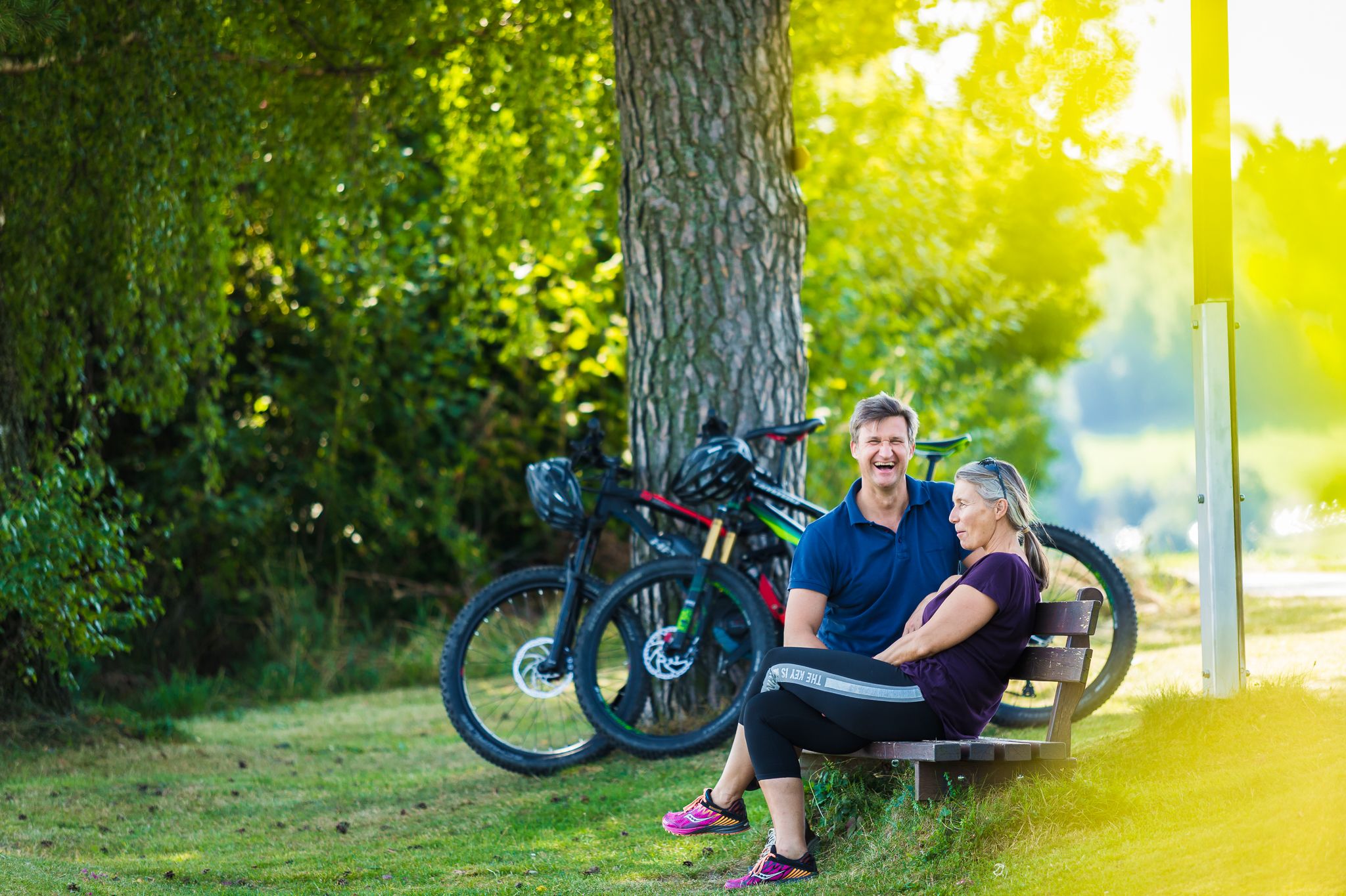 Ein Mann und eine Frau sitzen auf einer Bank im Grünen, zwei E-Bikes stehen daneben.