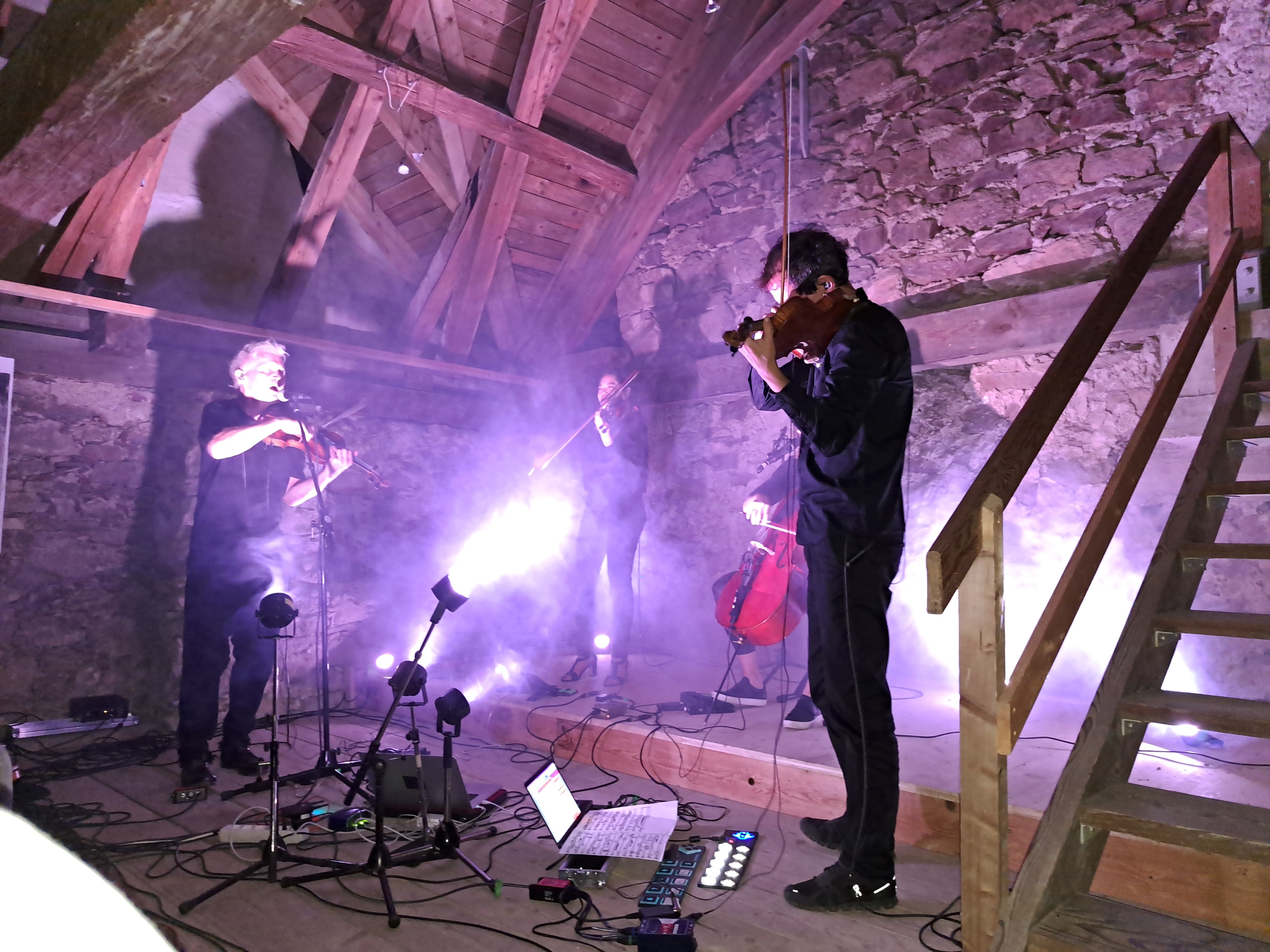 4 Musiker:innen mit Geigen und Kontrabass stehen in einem violett beleuchteten Raum. Scheinwerfer, Mikrofone und Laptops stehen bei den Musiker:innen. Der Raum besteht aus Holz und Stein, rechts im Bild ist eine Stiege aus Holz erkennbar.