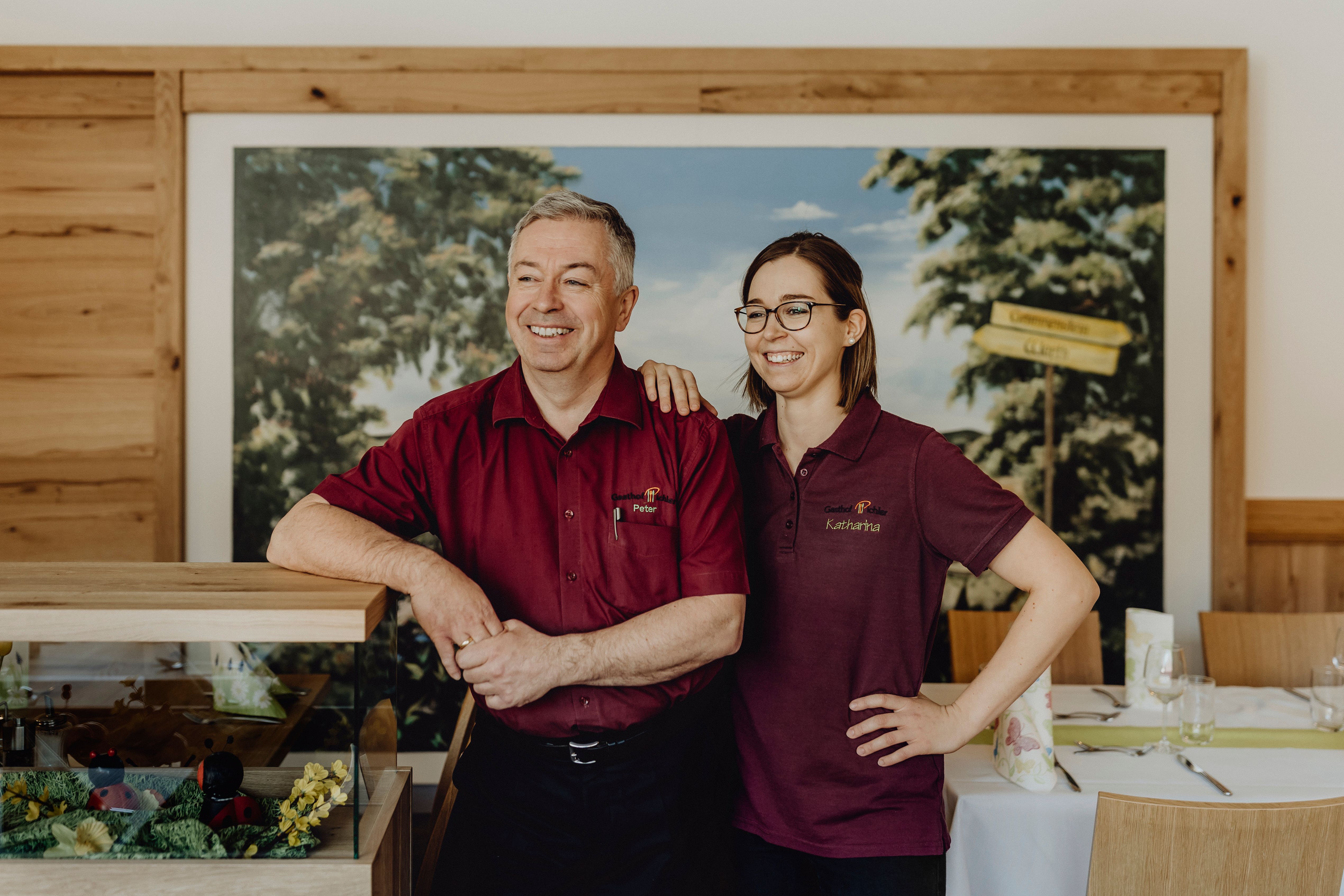 Zwei Personen in roten Hemden stehen lächelnd in einem Raum eines Gasthofes.