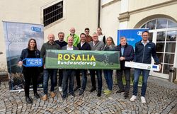 Mehrere Personen stehen in einem Innenhof und halten ein Wiener Alpen Schild, ein LEADER-Schild und ein Rosalia-Rundwanderweg-Schild in ihren Händen.