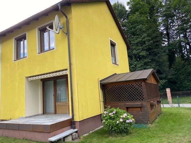 Gelbes Ferienhaus mit Terrasse und kleinem Anbau, umgeben von Bäumen.