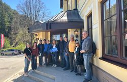 Eine Gruppe von rund 12 Personen steht auf den Stufen vor dem "Landgasthof zur Schubertlinde". Eine Frau hält ein Wiener Alpen Logo-Schild in der Hand.