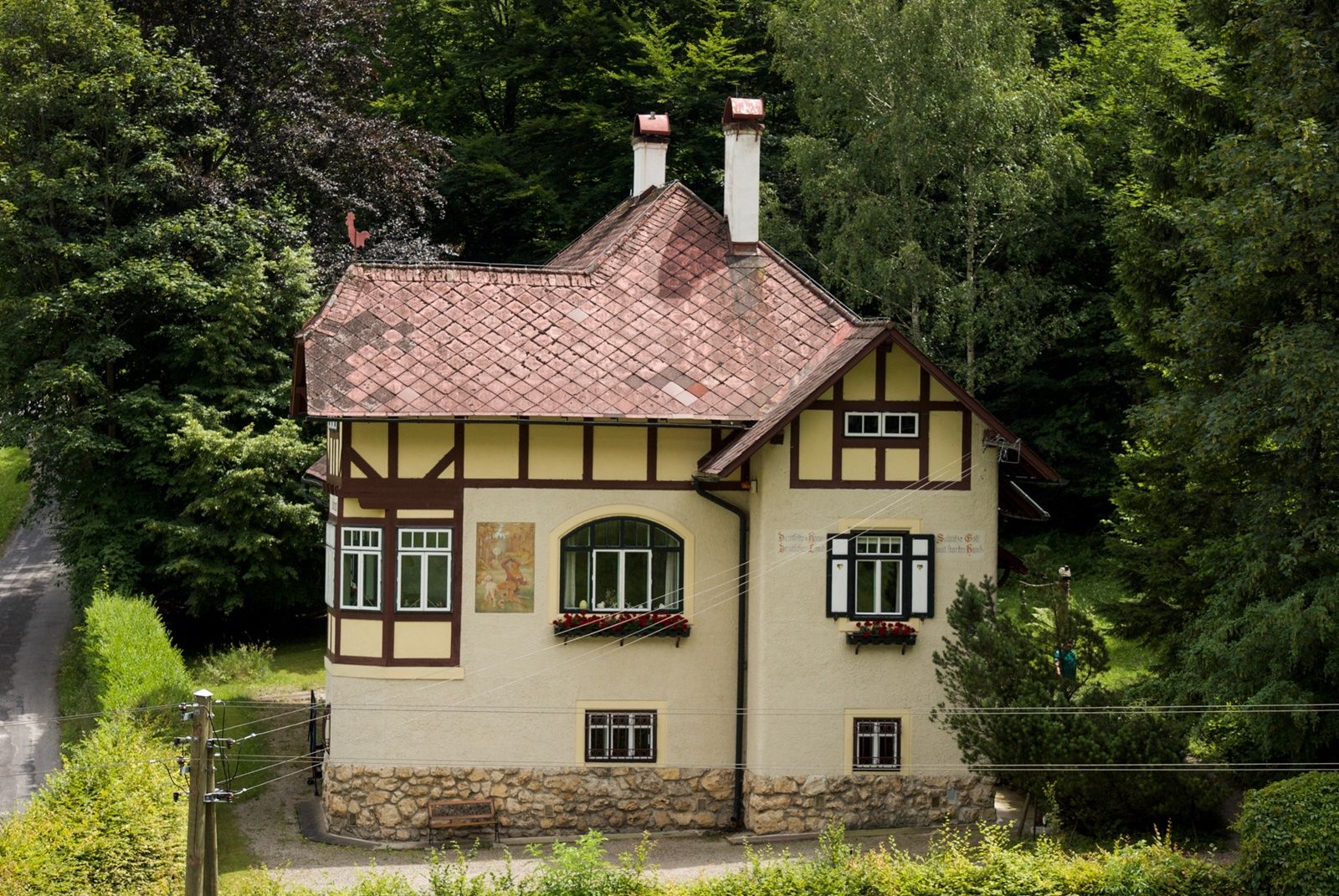 A two-storey house with half-timbering and red roof tiles, surrounded by trees.