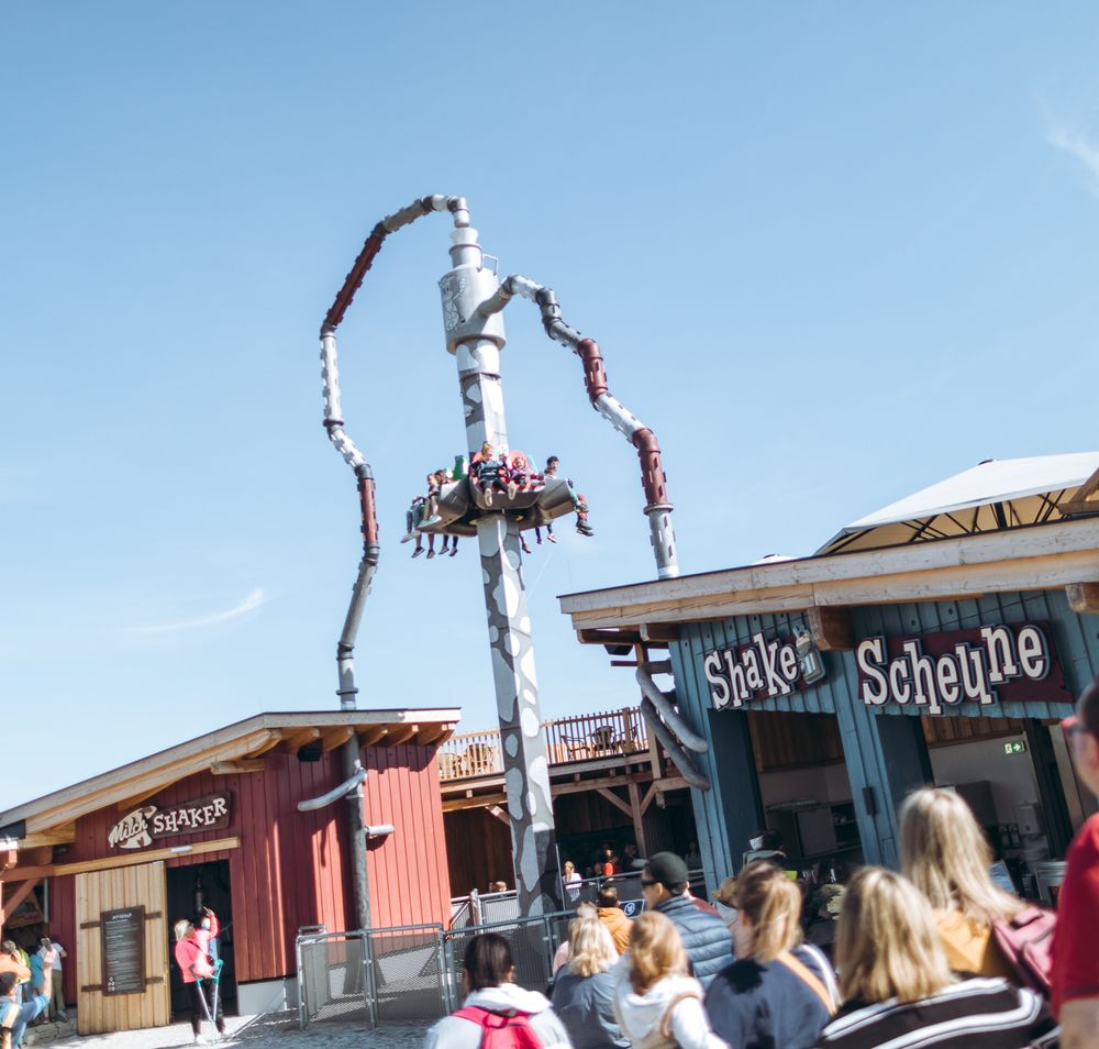 ein einige Meter hohes Fahrgeschäft mit zwei Sitzen an jeder Seite in denen Erwachsene und Kinder sitzen. Ganz oben ist eine große Milchkanne von der links und rechts kurvige Röhren weggehen. Im Vordergrund Menschen und ein rotes und ein blaues Gebäude aus Holz, mit den Aufschriften "Milch Shaker" und "Shake Scheune"