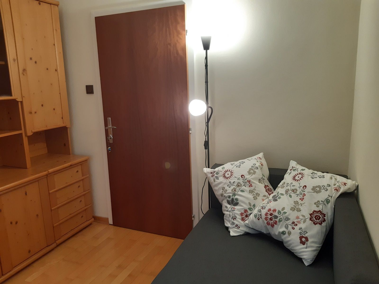 Ein gemütliches Zimmer mit einer grauen Couch, dekoriert mit zwei bestickten Kissen, einem Holzschrank und einer Stehlampe neben einer Holztür.