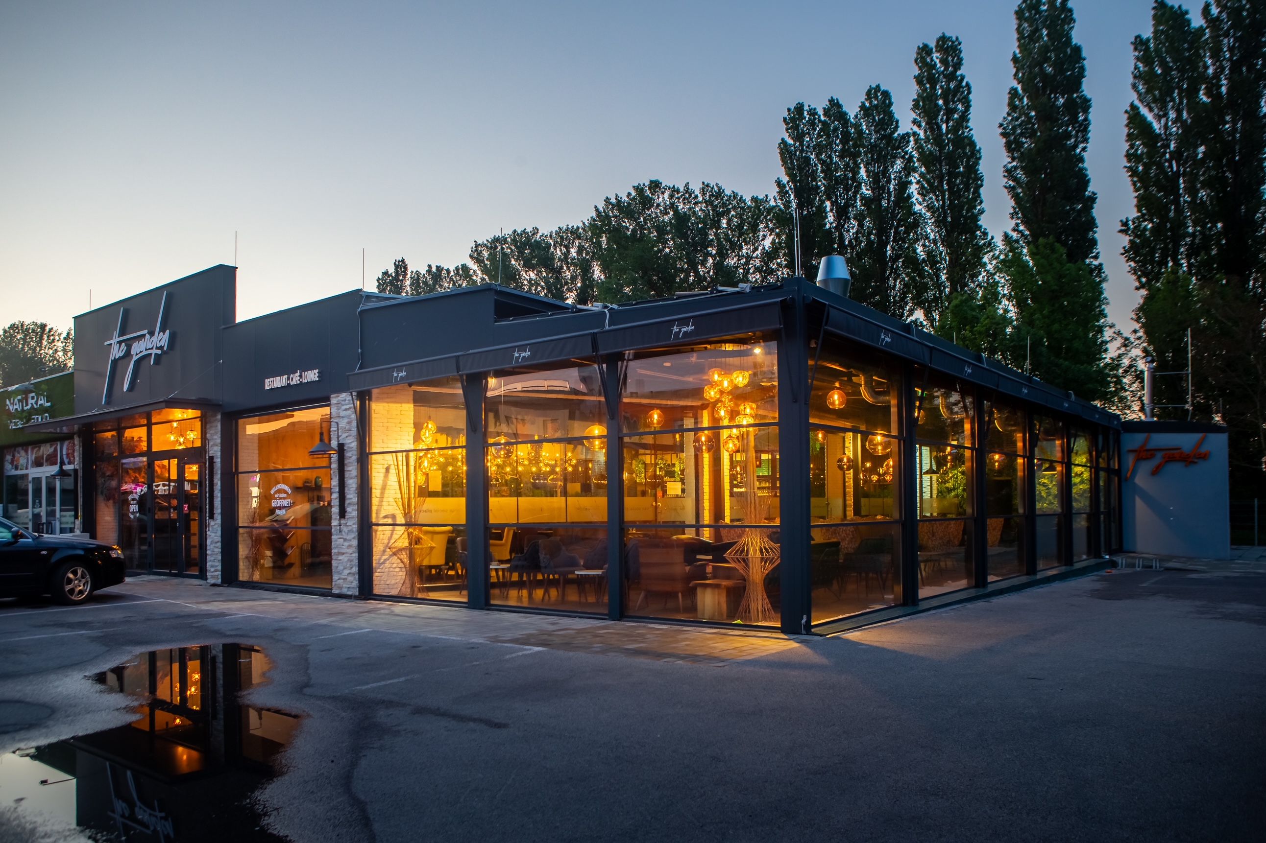 Außenansicht eines modernen Restaurants bei Dämmerung mit großen Glasfenstern und warmem Licht.