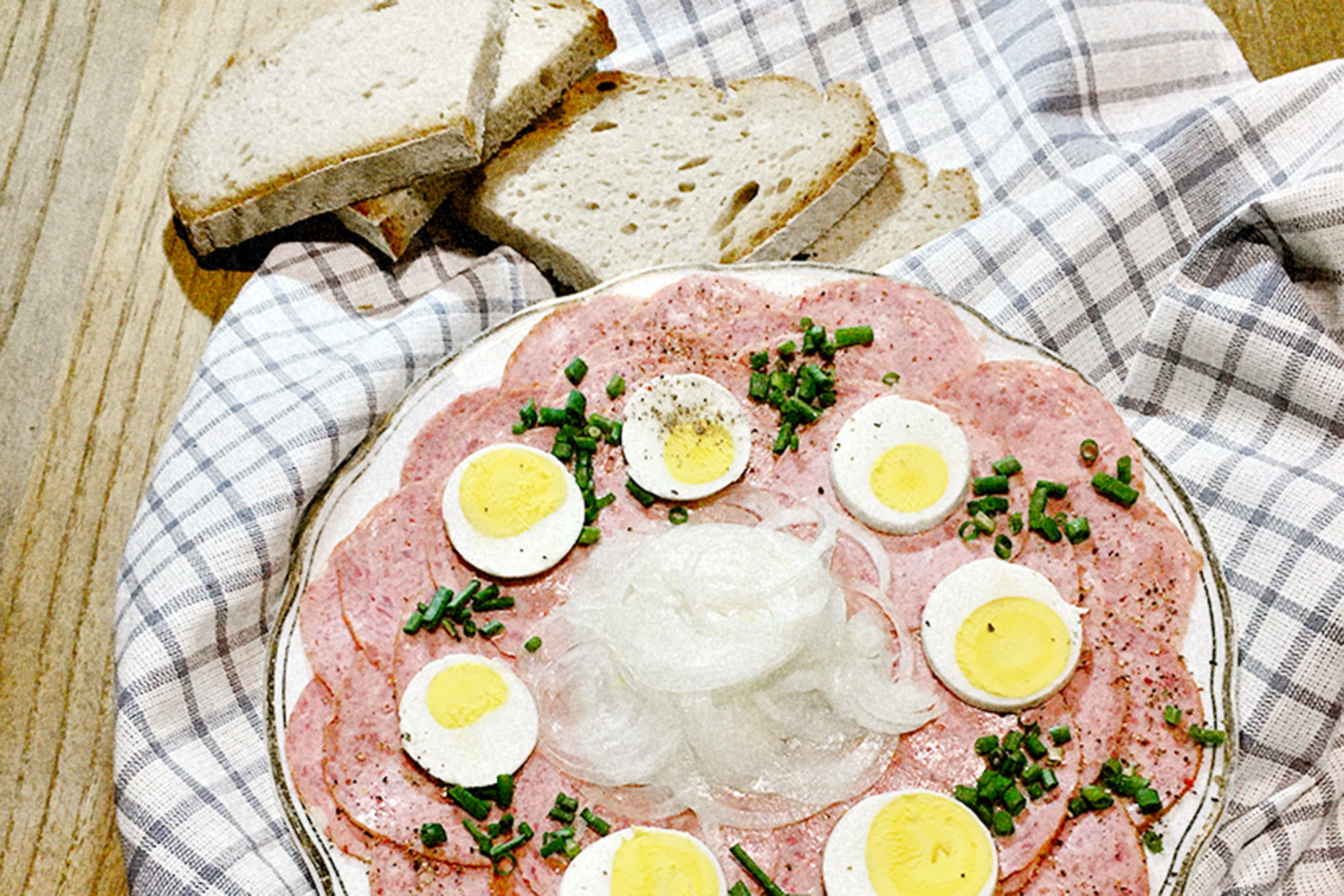 Ein Teller mit Wurstscheiben, gekochten Eiern und Zwiebeln, garniert mit Schnittlauch, neben Brotscheiben auf einem karierten Tuch.