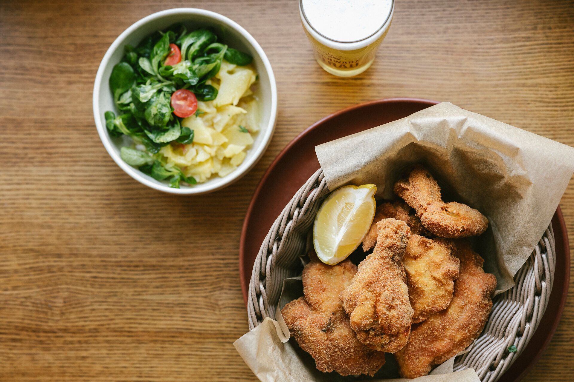 Korb mit knusprig panierten, frittierten Fleischstücken und Zitronenscheibe; im Hintergrund eine kleine Schüssel mit Salat und ein Glas Bier.
