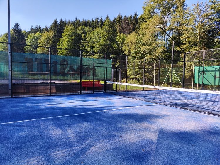 Padelplatz mit blauem Boden und Netz, umgeben von Bäumen.