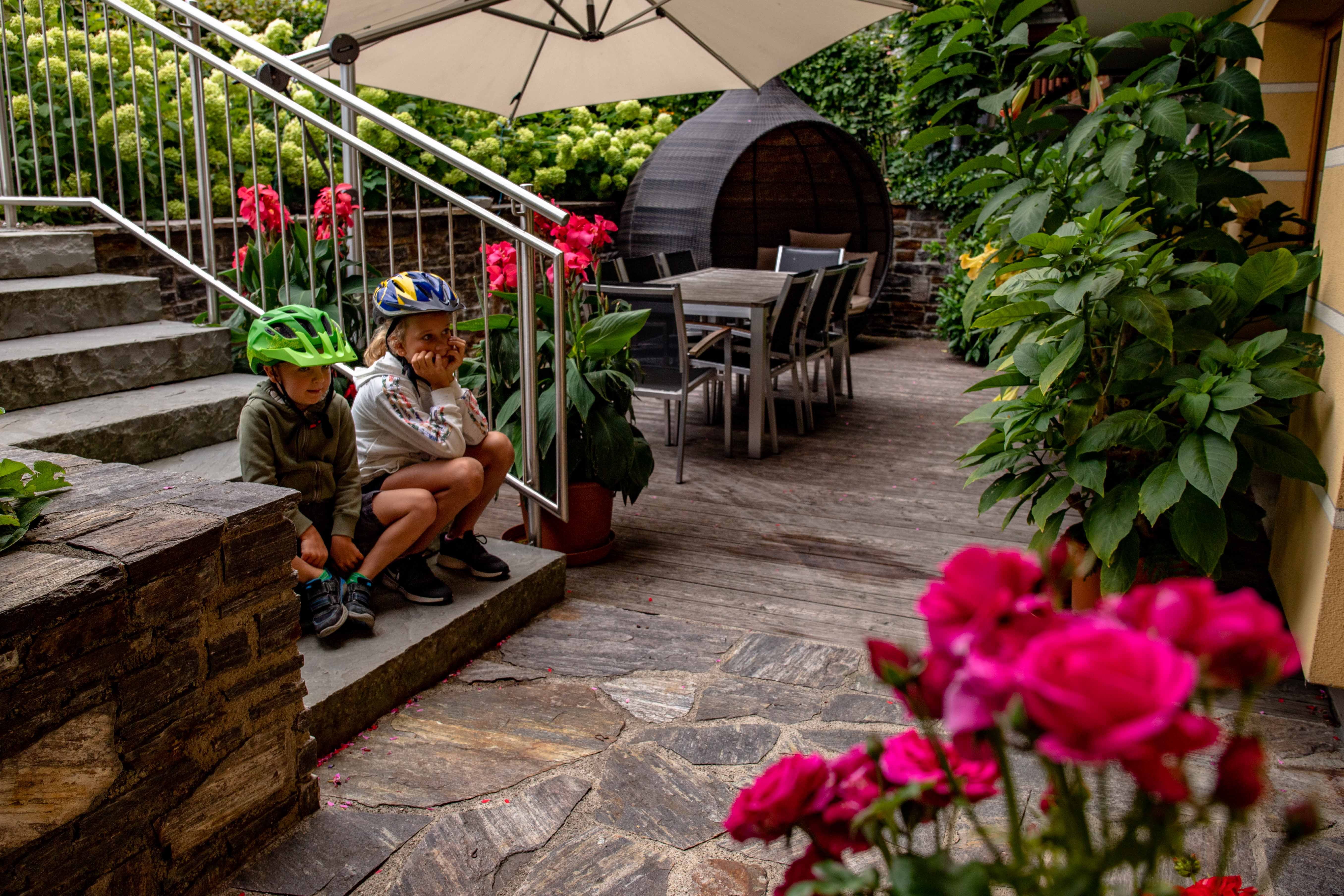 Zwei Kinder mit Fahrradhelmen sitzen auf einer Treppe in einem Garten mit Blumen und Gartenmöbeln.
