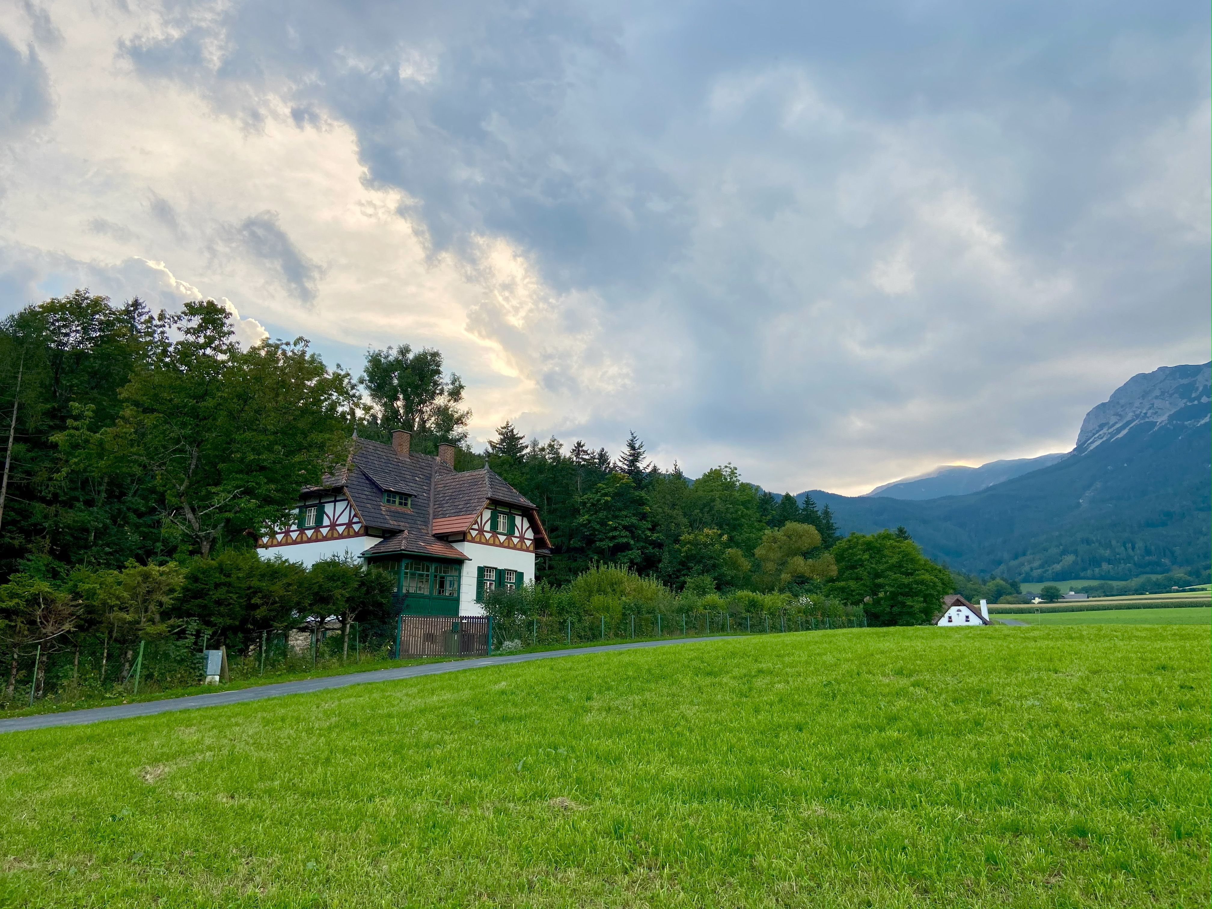 Ein Haus im traditionellen Stil steht vor einem bewaldeten Hügel, umgeben von grünen Wiesen und Bergen im Hintergrund.