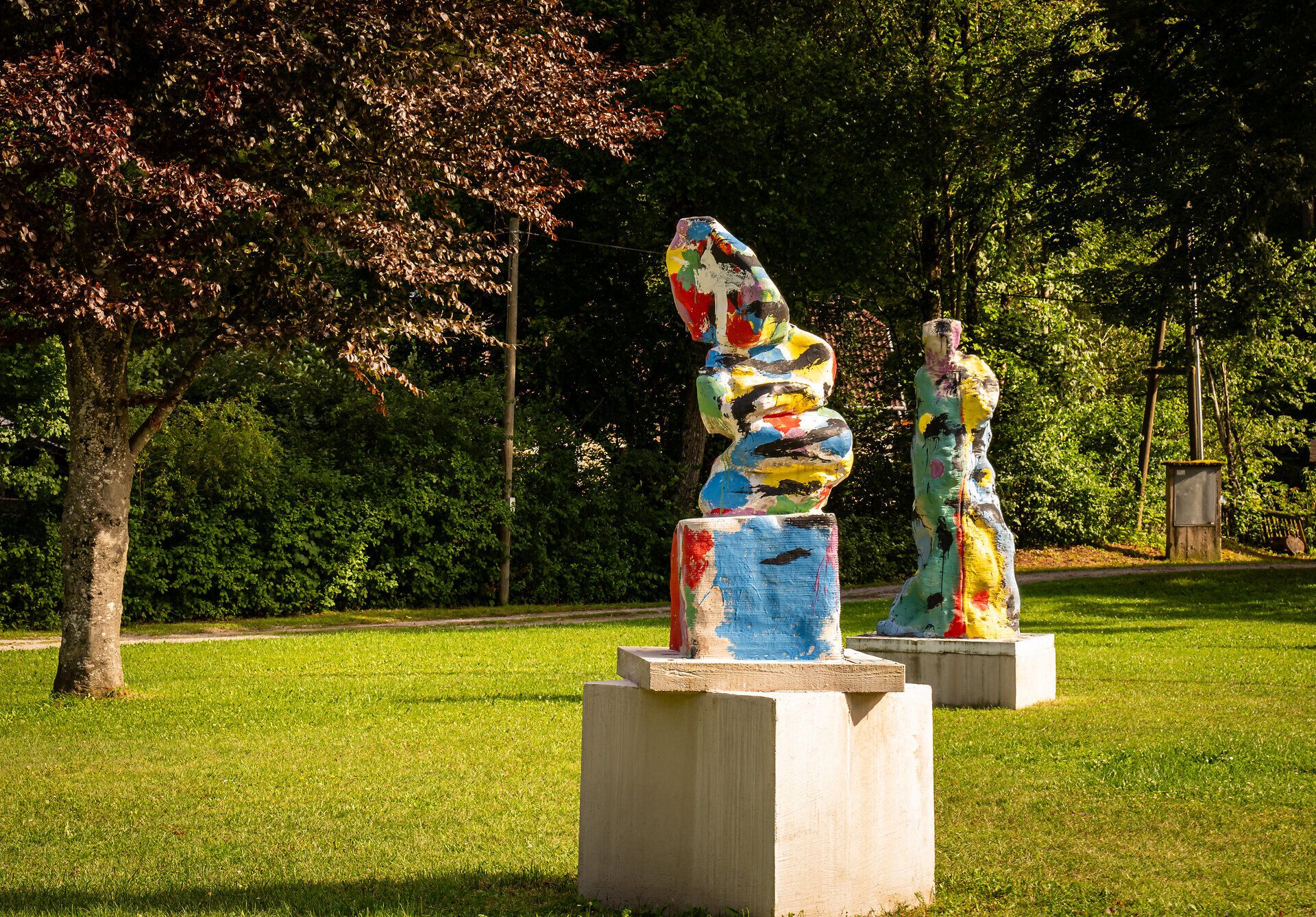 Bunte Kunstwerke stehen auf quadratischen Sockeln auf einer grünen Wiese.