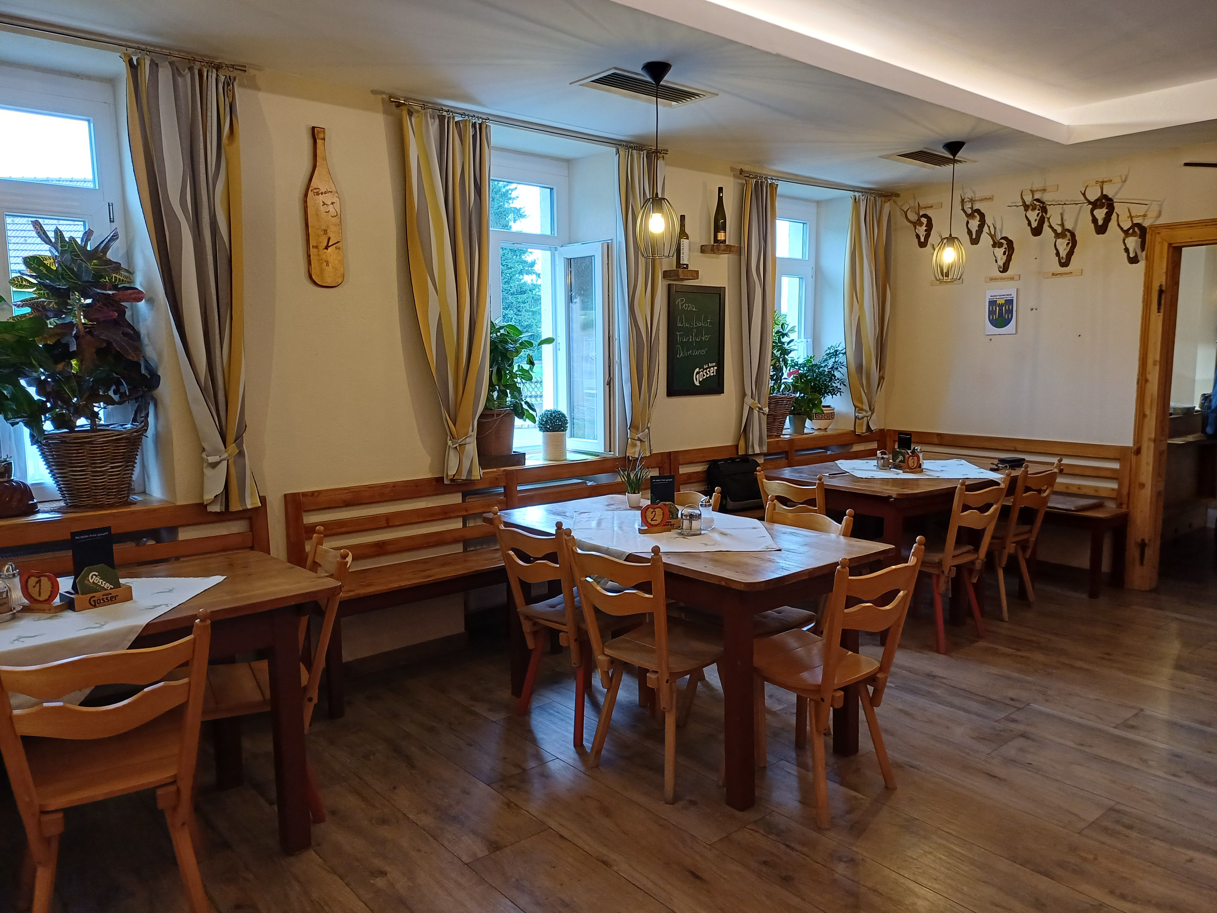 Innenansicht eines gemütlichen Gastraums mit Holztischen und Stühlen, dekoriert mit Pflanzen und Hirschgeweihen an der Wand.