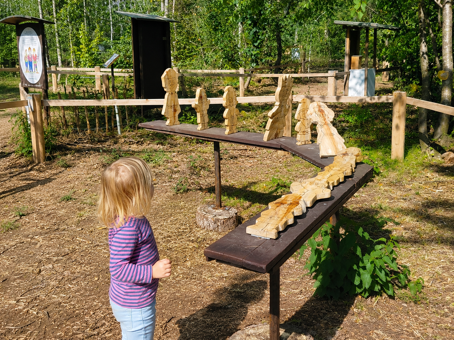 Ein Kind betrachtet eine Reihe von Holzfiguren in einem Märchenwald. Die Figuren sind auf einem Tisch im Freien aufgestellt.