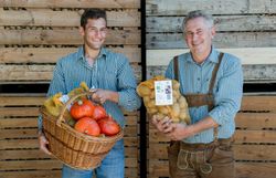 Zwei Männer vor einer Holzwand, einer hält Kartoffeln, der andere einen Korb mit Kürbissen.