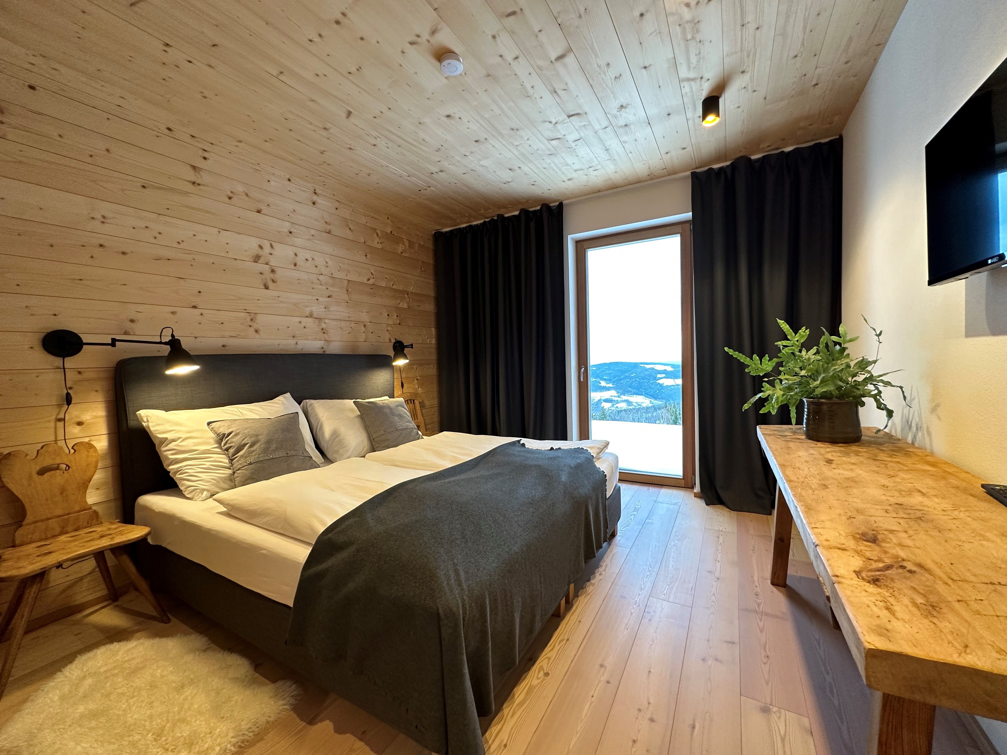 Gemütliches Schlafzimmer mit Holzwänden, Doppelbett, Holzstuhl, Tisch mit Pflanze und großem Fenster mit Vorhängen.