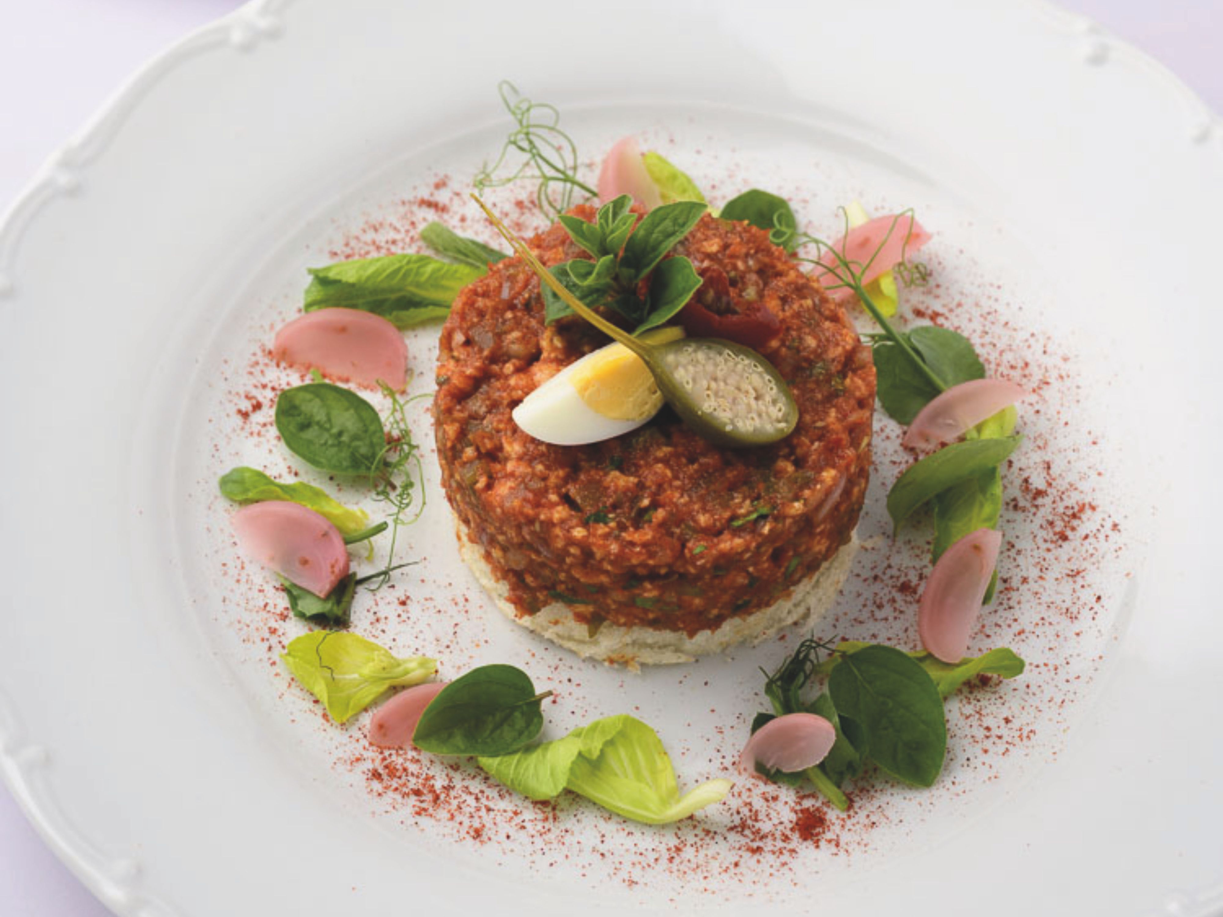 Ein kunstvoll angerichtetes Beef Tartar auf einem weißen Teller, bestehend aus einer runden Portion Tatar, garniert mit Kapern, Eierscheiben und frischen Kräutern.