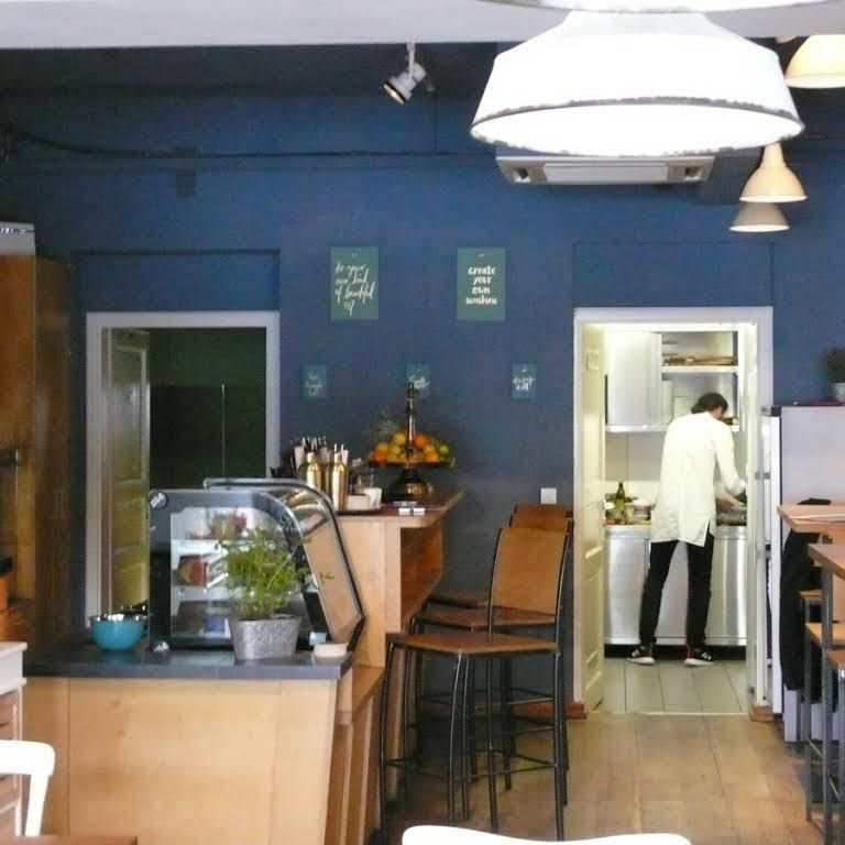 Innenansicht eines Cafés mit blauer Wand, Theke, Barhockern und einer Person in der Küche.