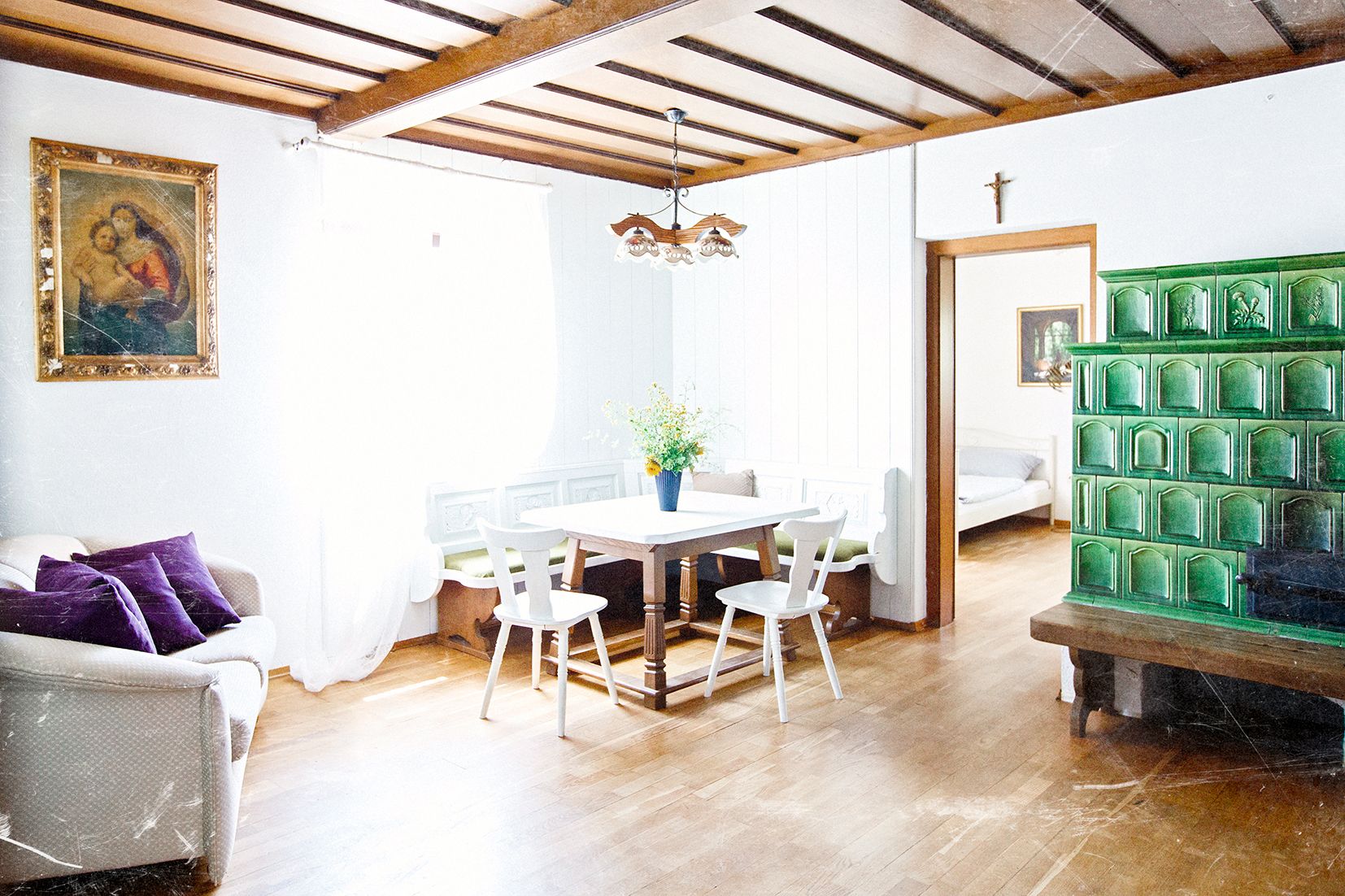Helles Wohnzimmer mit Holzbalkendecke, grüner Kachelofen, Esstisch mit Stühlen und Sofa. An der Wand hängt ein Gemälde.