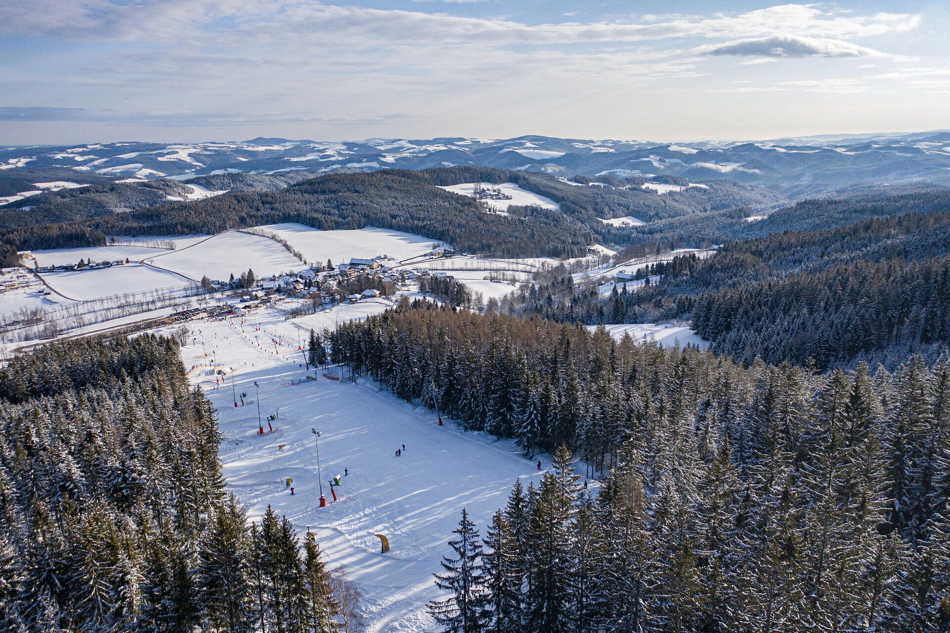 Blick über das verschneite Kinderskiland der Wexl Arena mit Skipisten, Liften und umliegenden Wäldern
