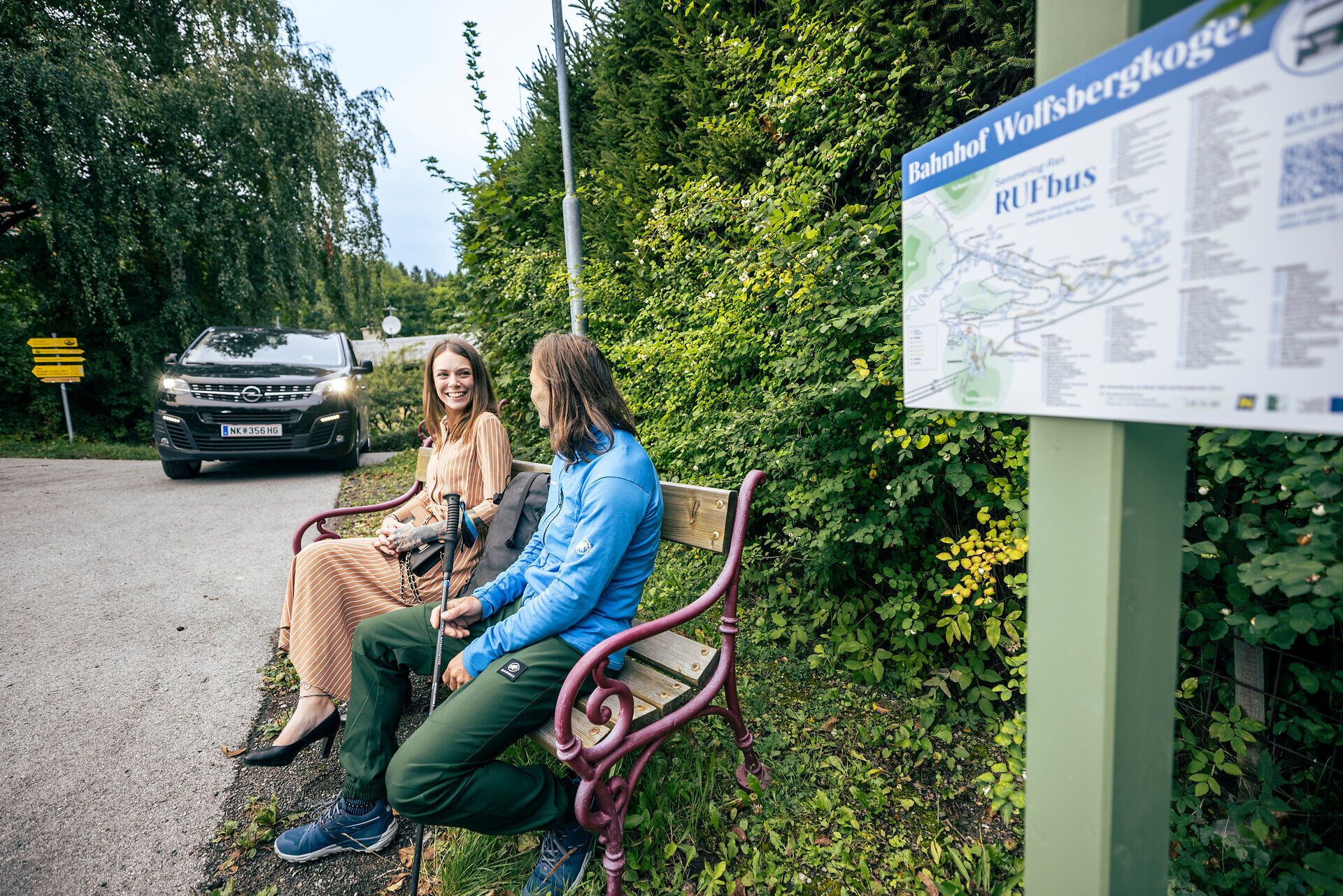 Ein schwarzer Kleinbus, der RUF Bus, holt gerade eine Frau in einem schönen orangen Kleid und einen Wanderer ab. 