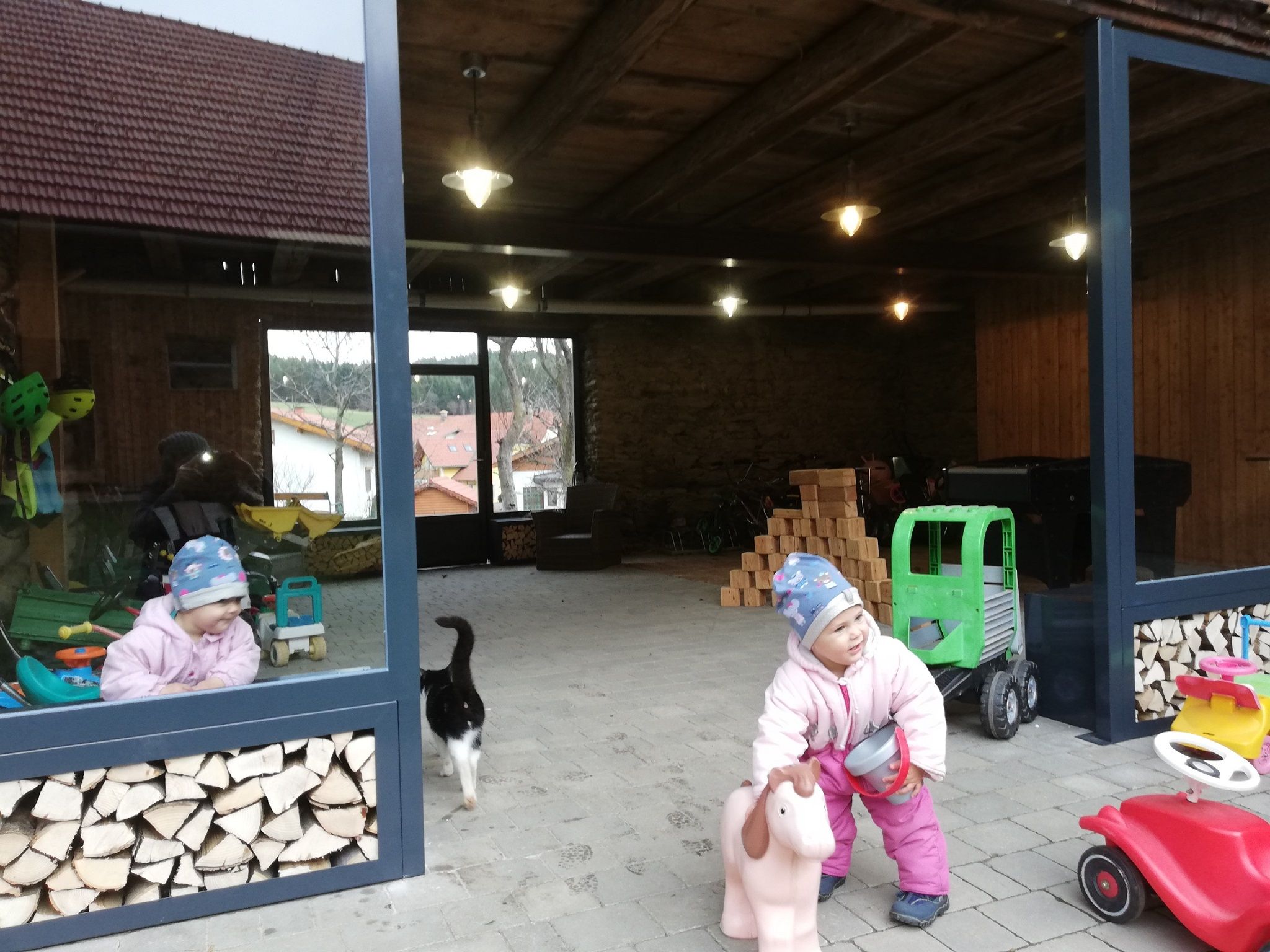 Ein Kind in Winterkleidung spielt mit einem Spielzeugpferd in einem offenen Raum mit Holzstapeln und Spielzeug. Eine Katze läuft vorbei.