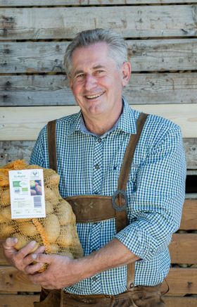 Ein Mann in Tracht gekleidet steht vor einer Holzwand und hält einen Sack mit Kartoffeln.