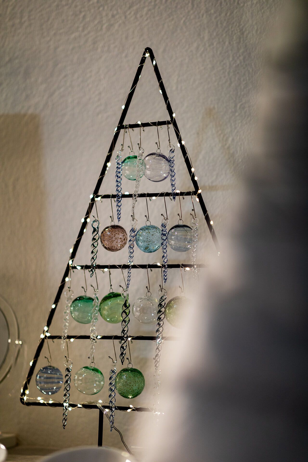 Minimalistischer Weihnachtsbaum aus Metall mit hängenden Glaskugeln an einer Wand.